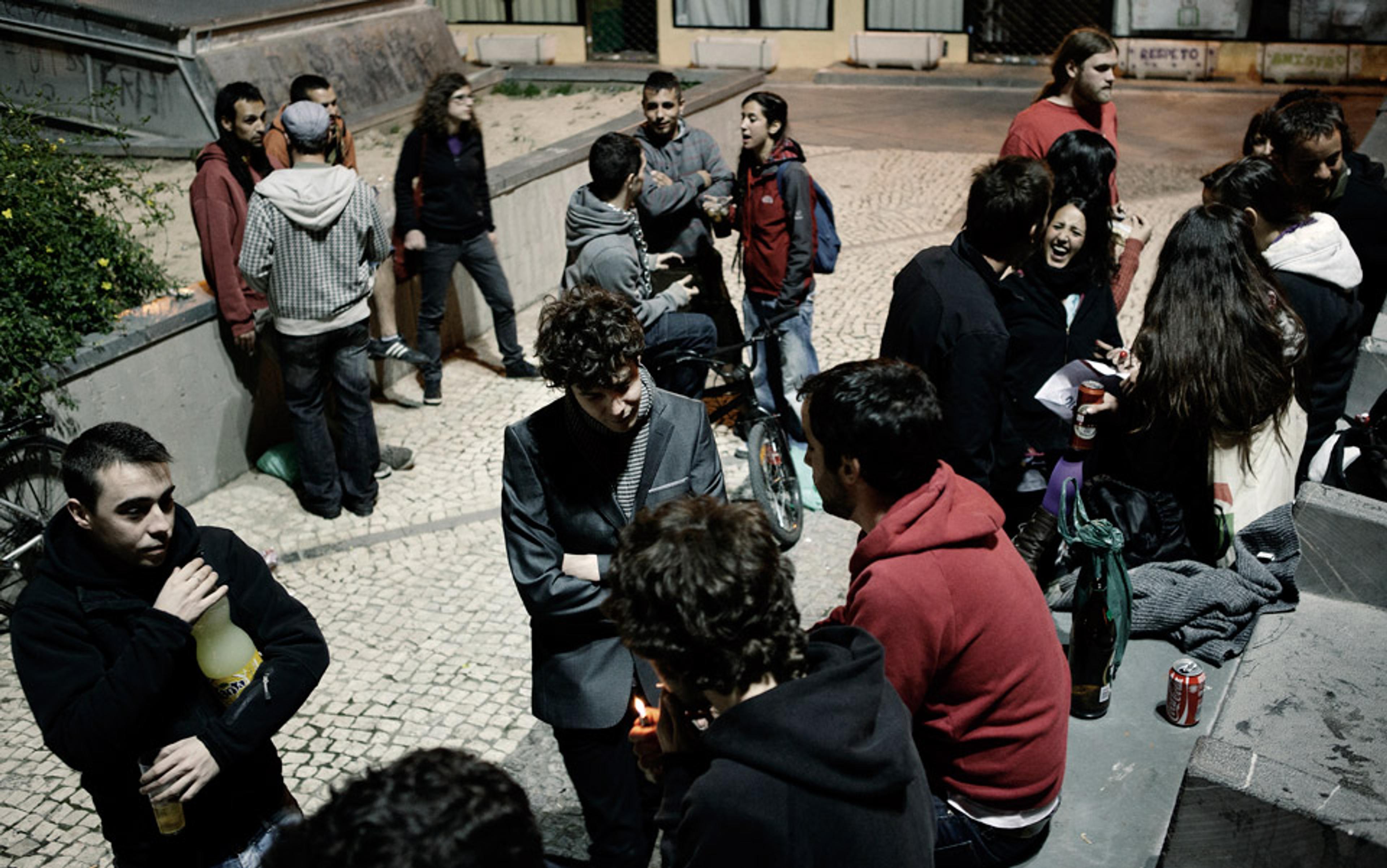 A group of people socialising outdoors at night, some holding drinks, gathered near steps and a stone wall in a city square.