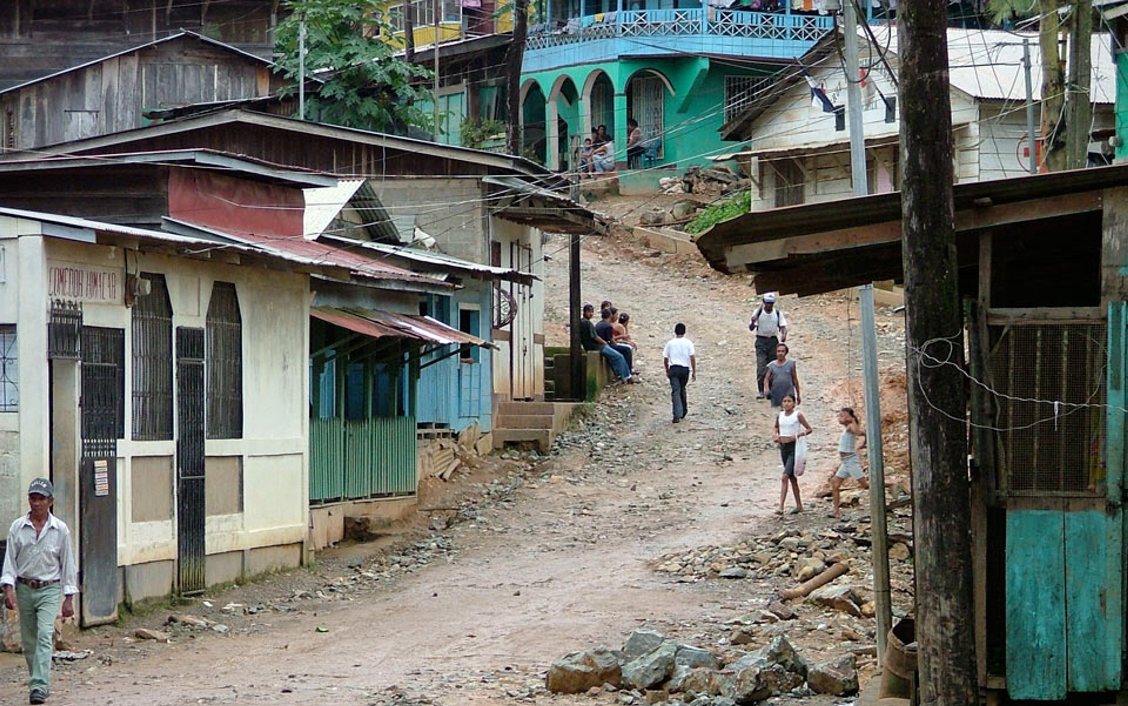 Photo of a rustic village street with people walking and buildings on both sides. The road is unpaved, and houses are of simple construction.