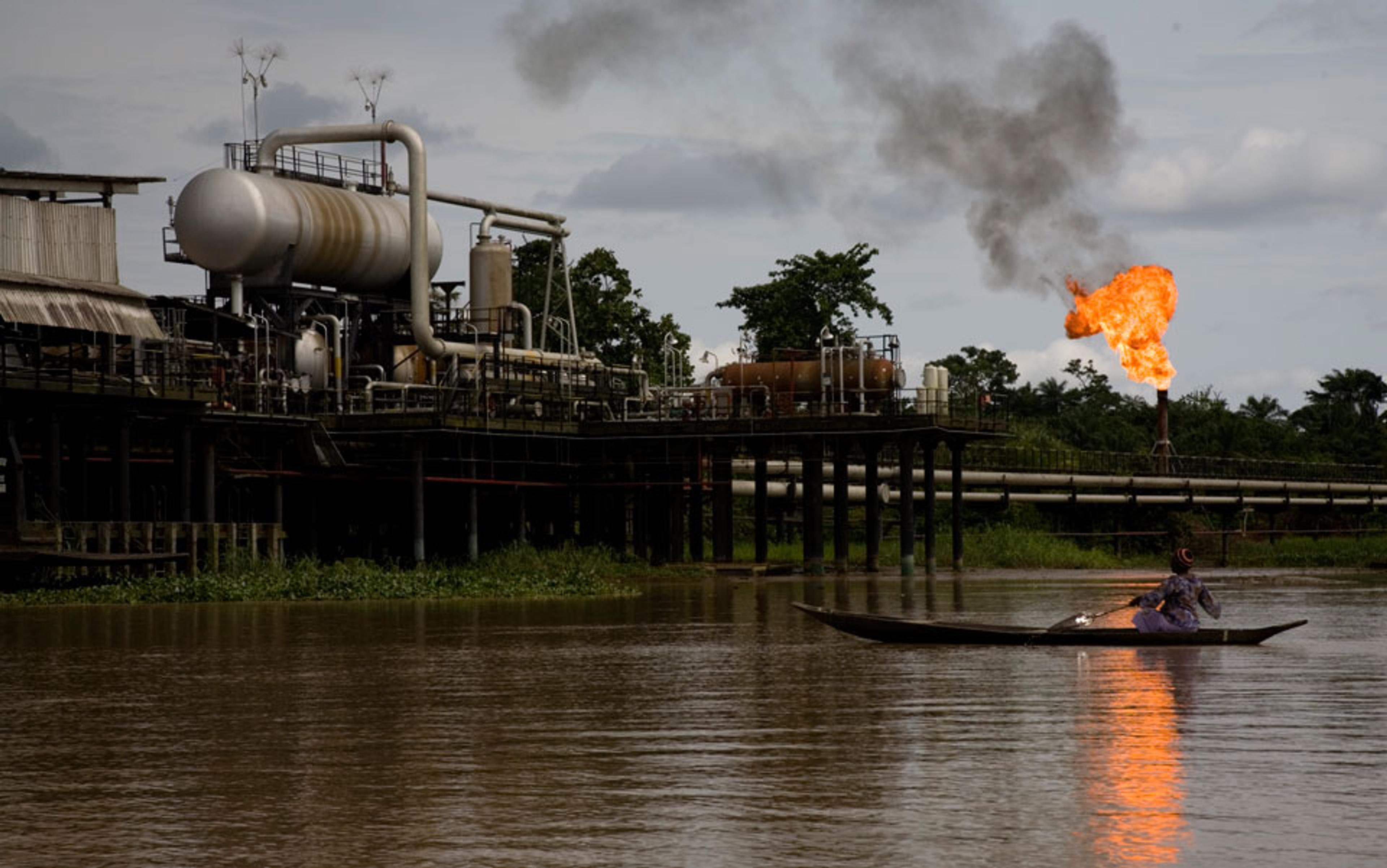 An oil refinery by a river with a person in a canoe in the foreground and a gas flare burning in the background.