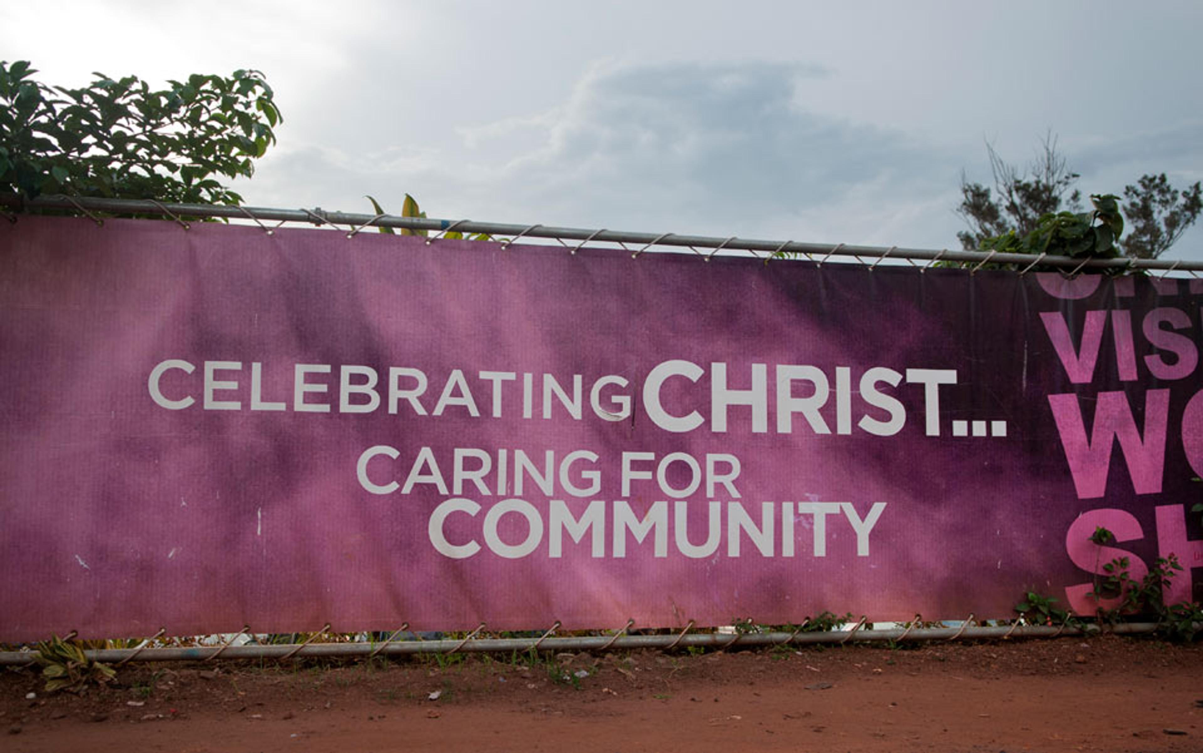 A purple banner on a fence reading “Celebrating Christ… Caring for Community” with foliage and a cloudy sky in the background.