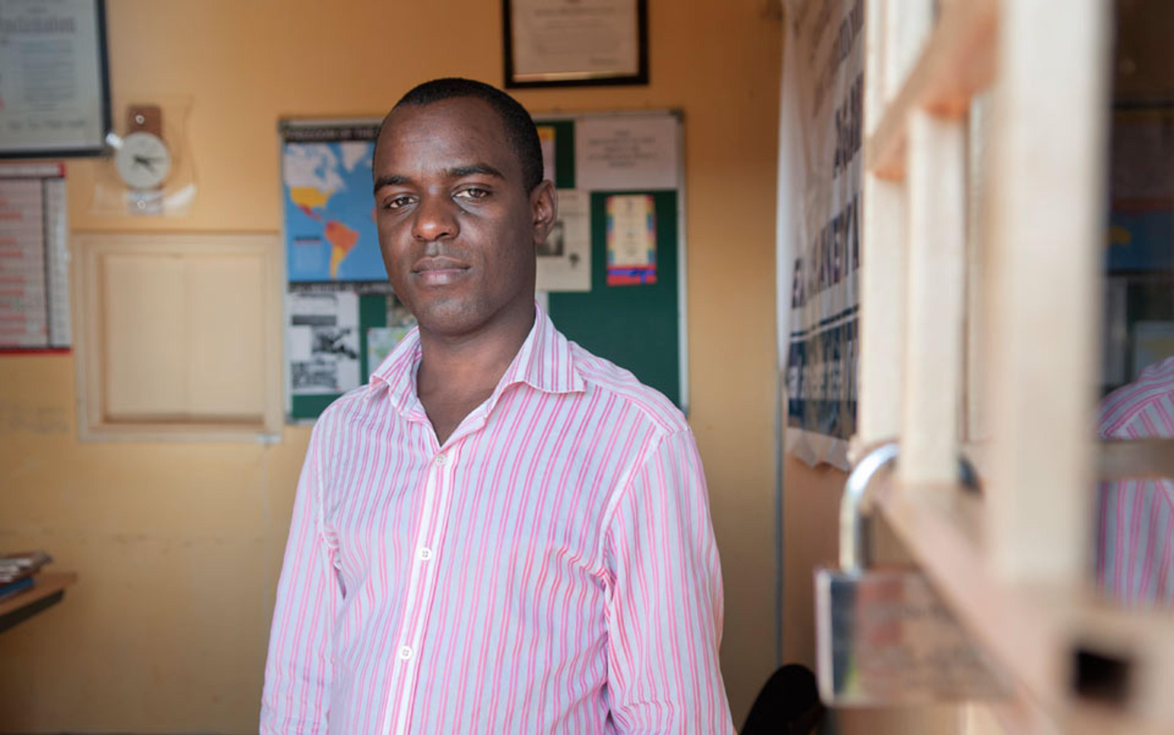 A Black man in a pink striped shirt standing in an office with a world map, certificates, and a clock on the wall.