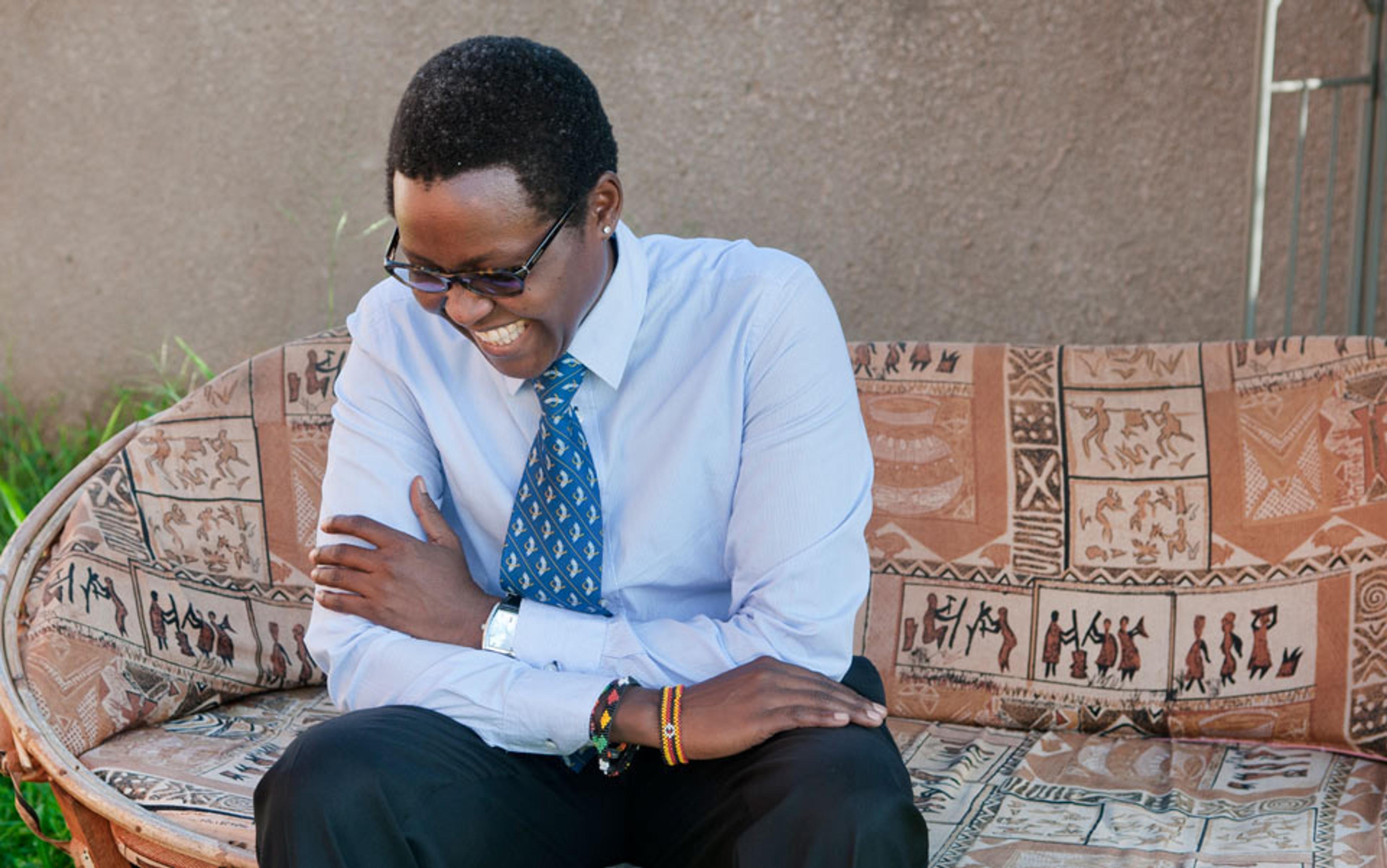 A Black man wearing glasses, a light blue shirt and a blue tie, smiling while sitting on a patterned couch outdoors.