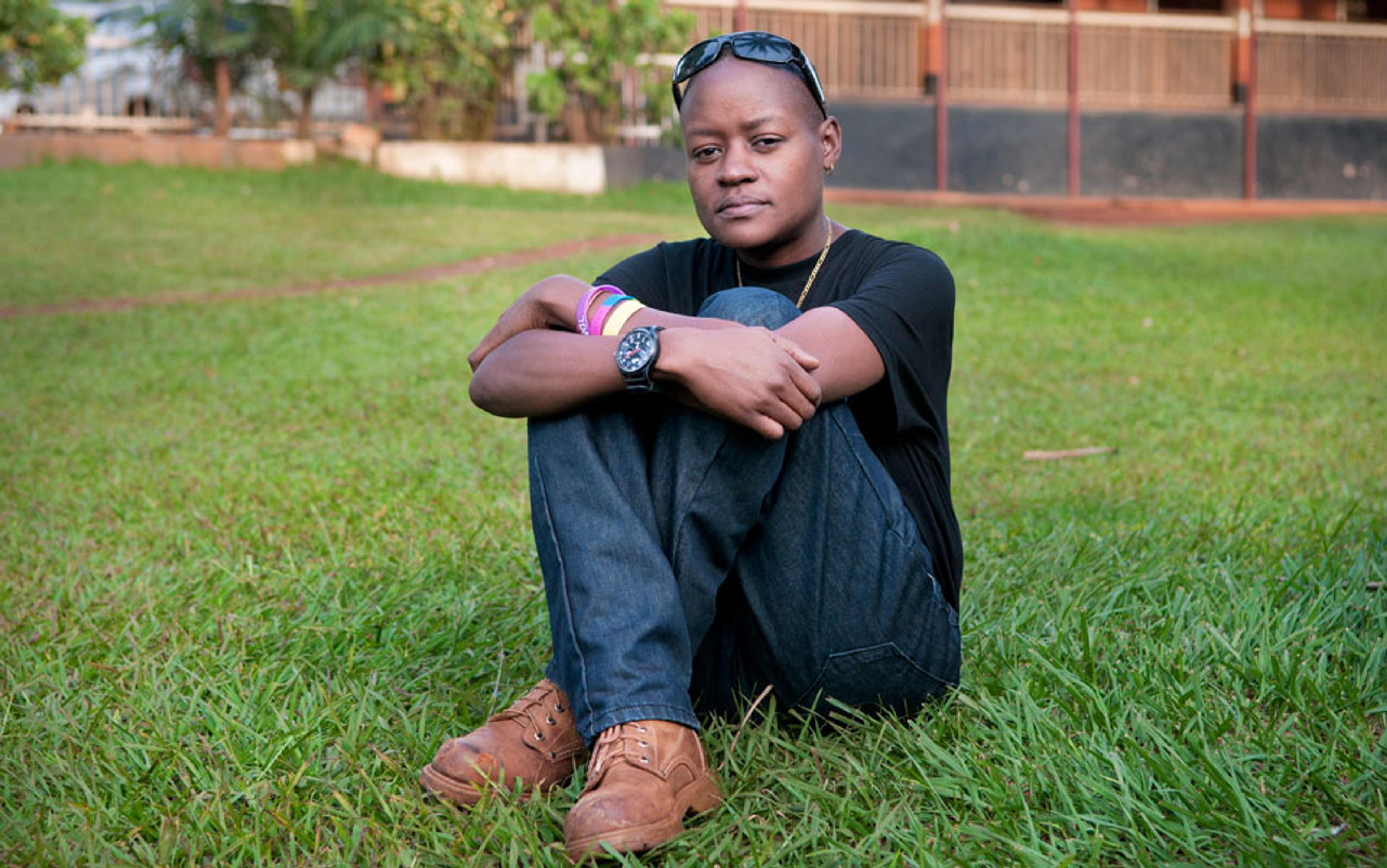 A Black man sitting on grass, legs crossed, wearing a black shirt, jeans and brown shoes with a building in the background.