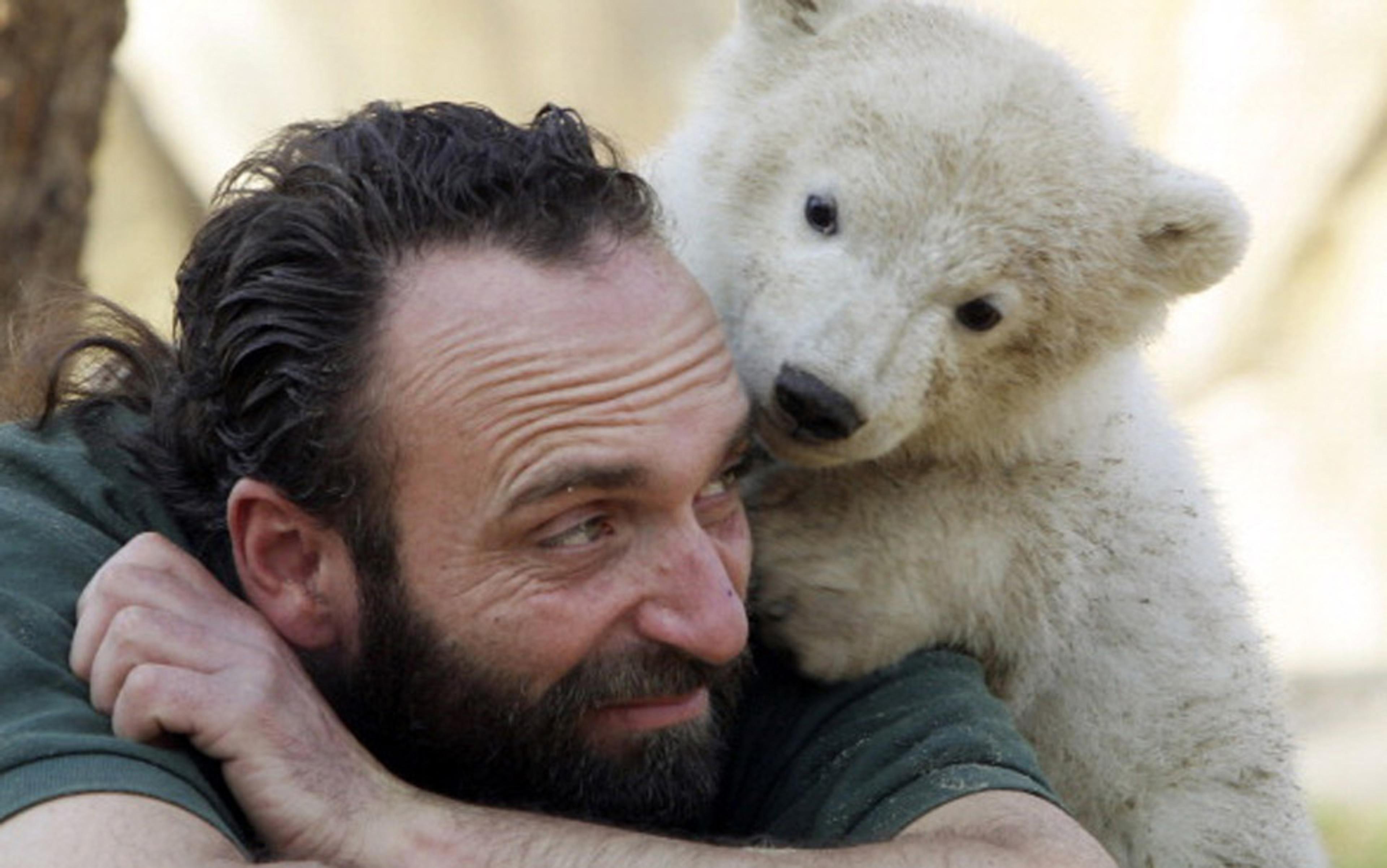 Photo of a smiling man with a beard cuddling a polar bear cub. The cub rests its head on the man’s shoulder while he looks at it.