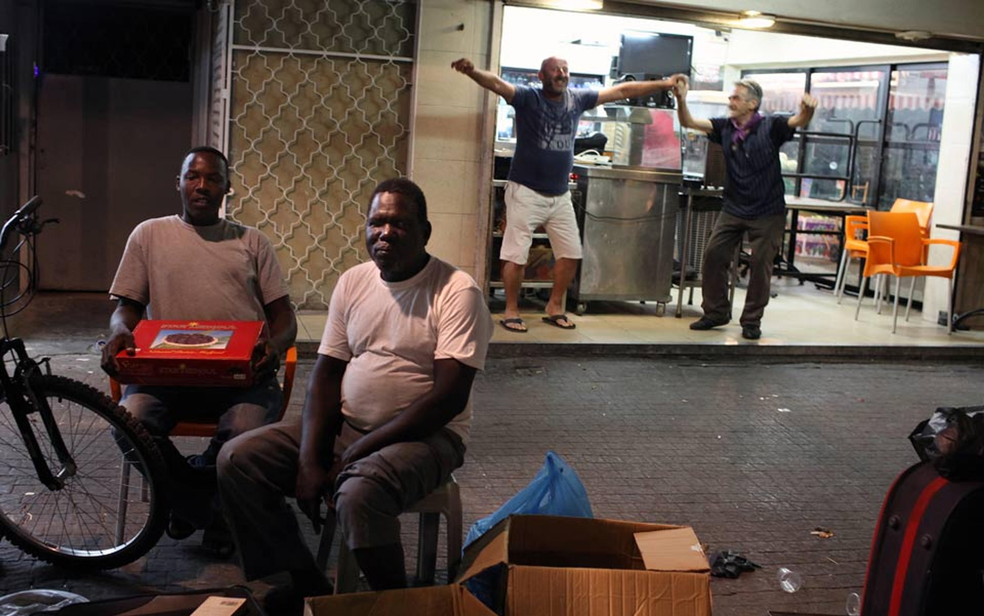 Two African men sit in front of a building, while two Israeli men stand behind them, smiling and holding hands in a dance.