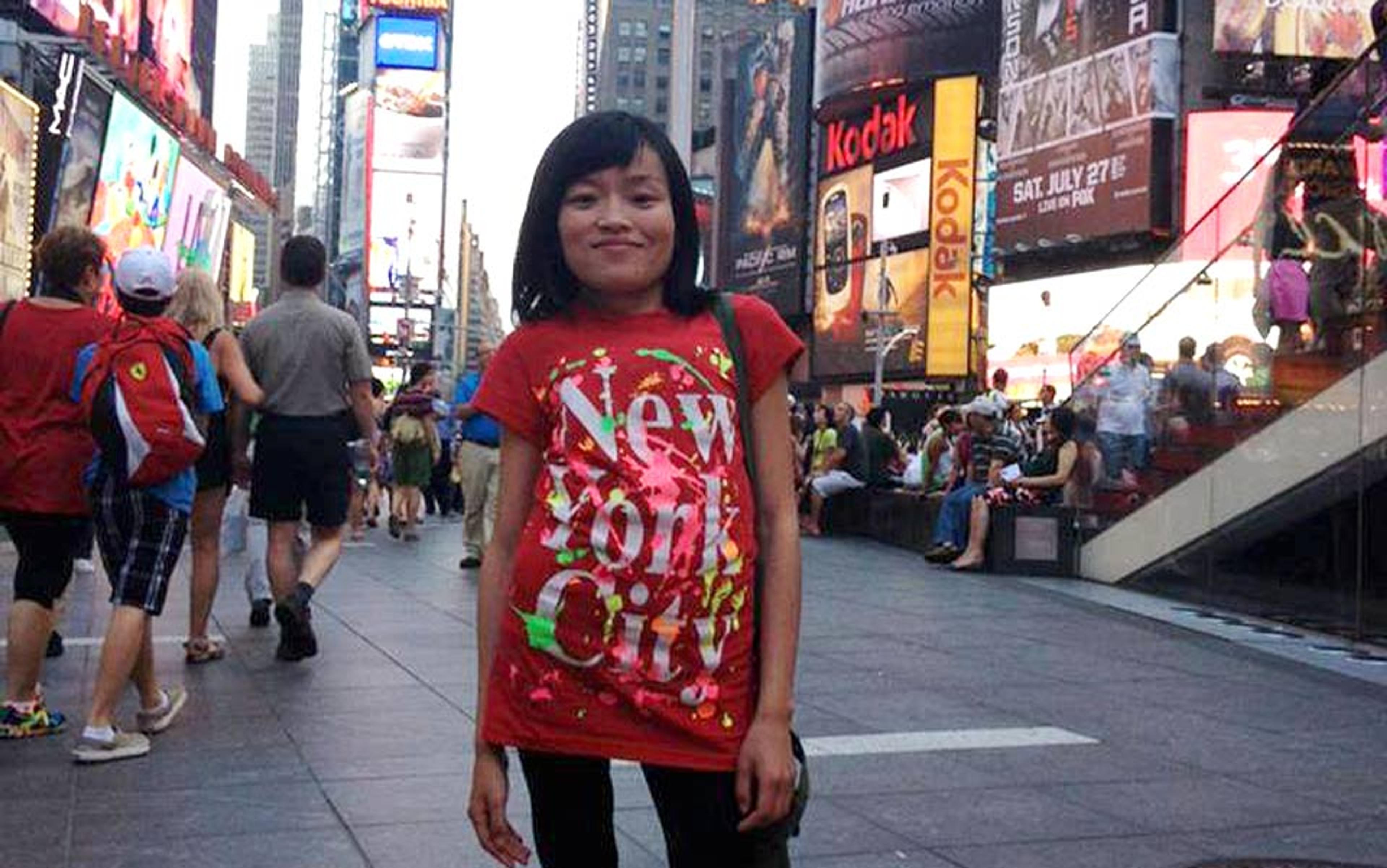 A smiling woman with scoliosis in a red “New York City” shirt standing in a bustling Times Square surrounded by people and bright billboards.