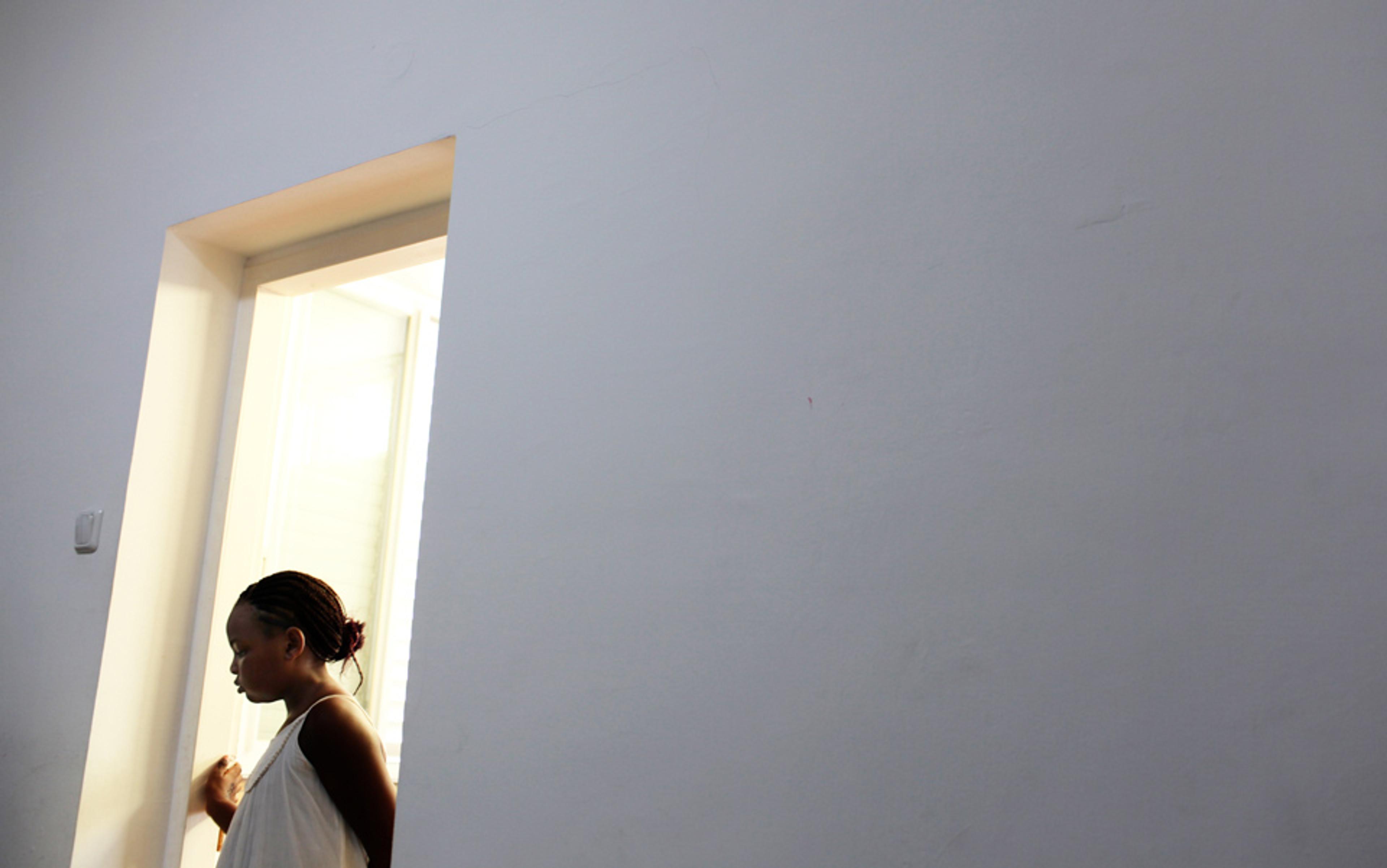 A Sudanese girl standing in a brightly lit doorway looking down, surrounded by a plain white wall with a minimalist aesthetic.