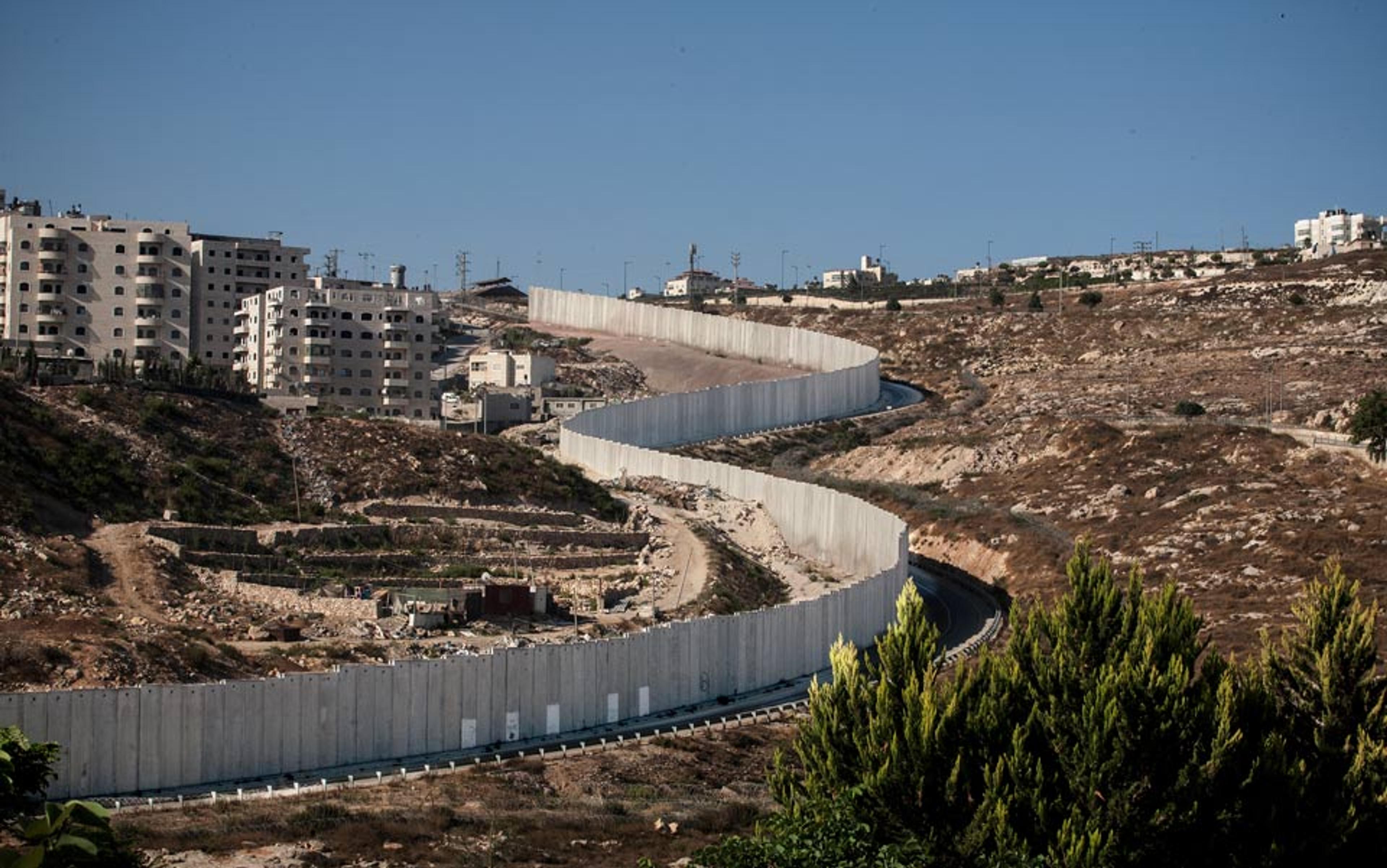 A long, winding concrete wall separating urban buildings from a rocky, barren landscape, under a clear blue sky.