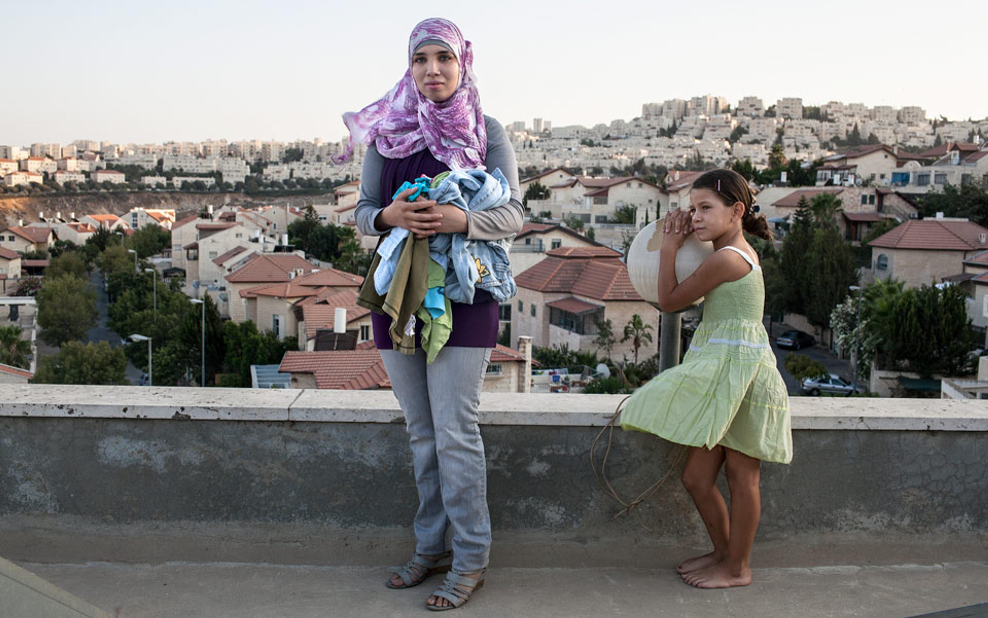 A woman in a headscarf holding laundry and a girl in a green dress overlooking a residential area with houses and trees.