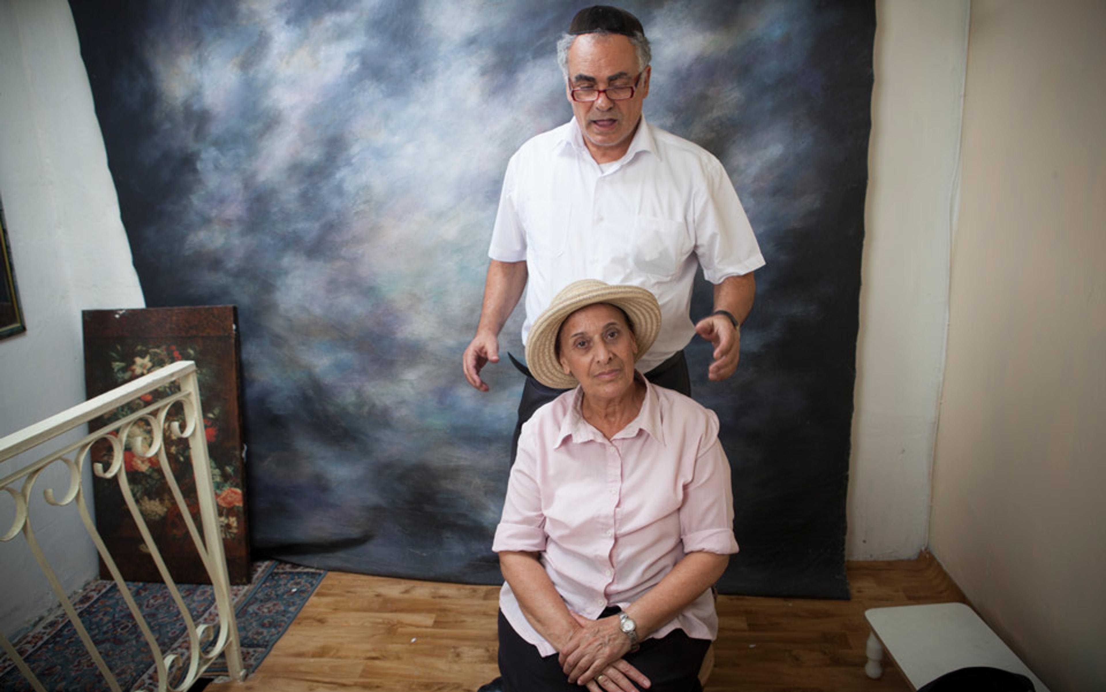 An elderly woman sitting and wearing a straw hat, with an elderly man in a kippah standing behind her in a room with a painted backdrop.