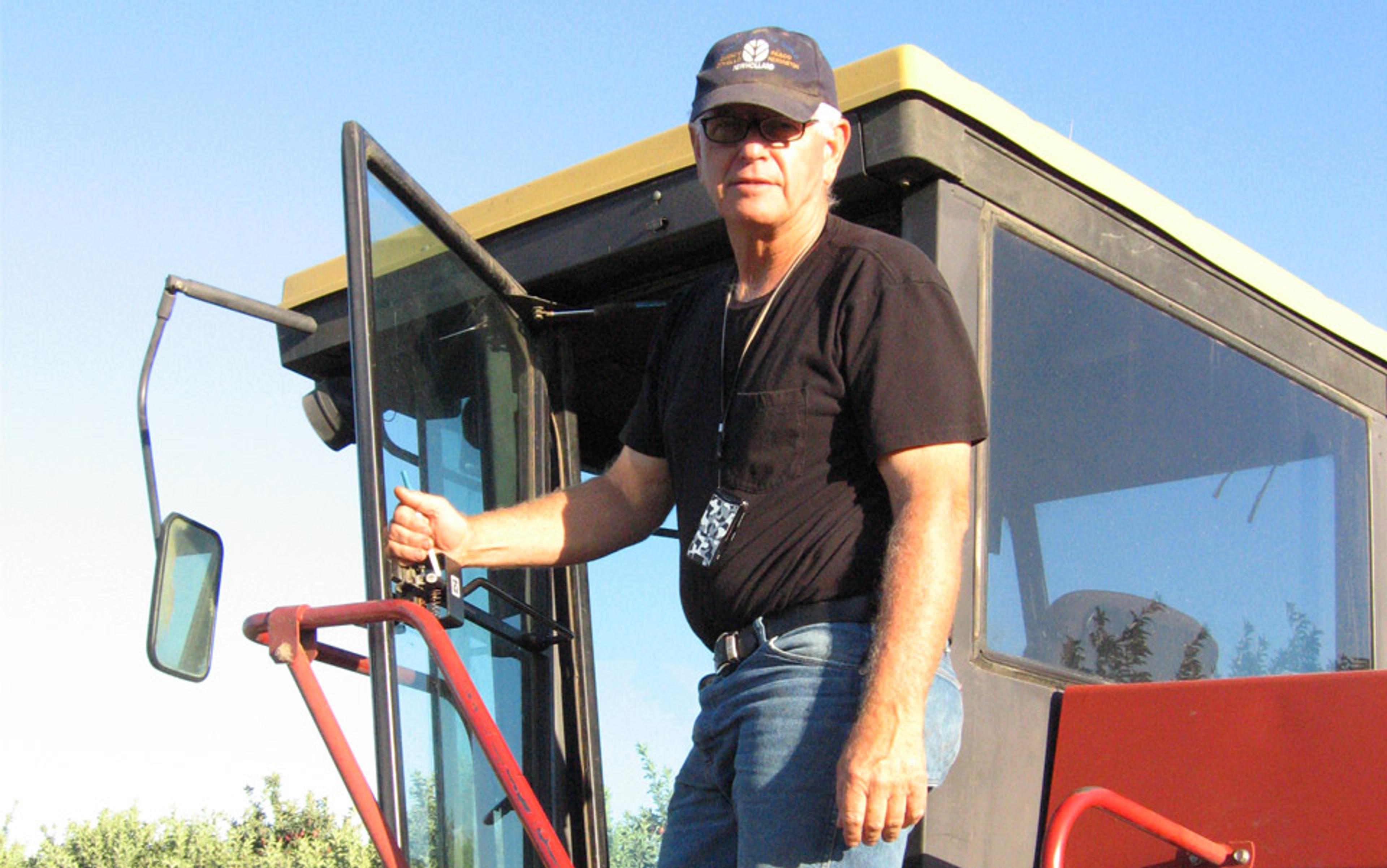 Photo of a man in a cap and black T-shirt standing on the steps of a tractor with a clear sky background.