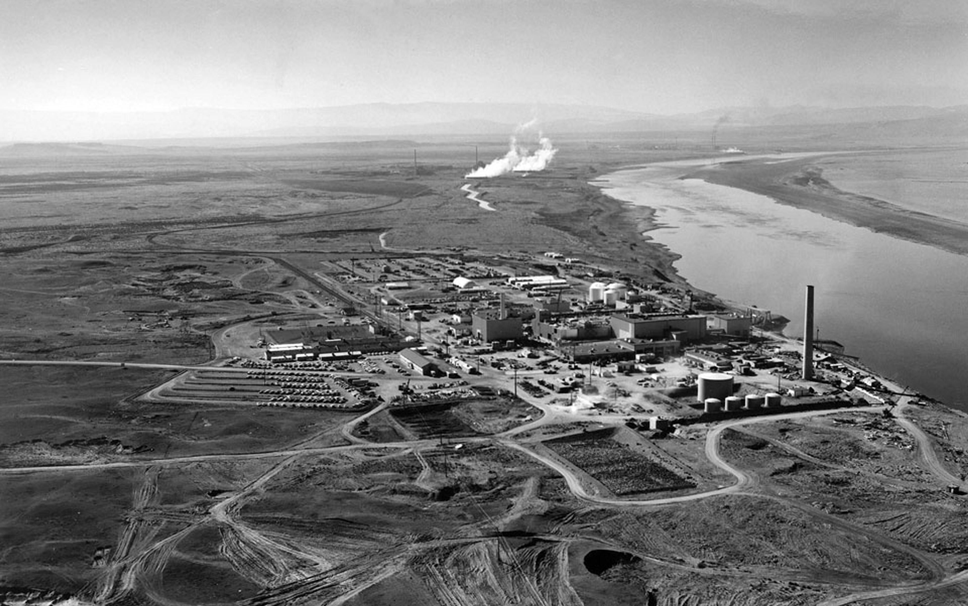 Aerial black and white photo of an industrial complex with smokestacks by a river in a barren landscape; vehicles parked nearby.