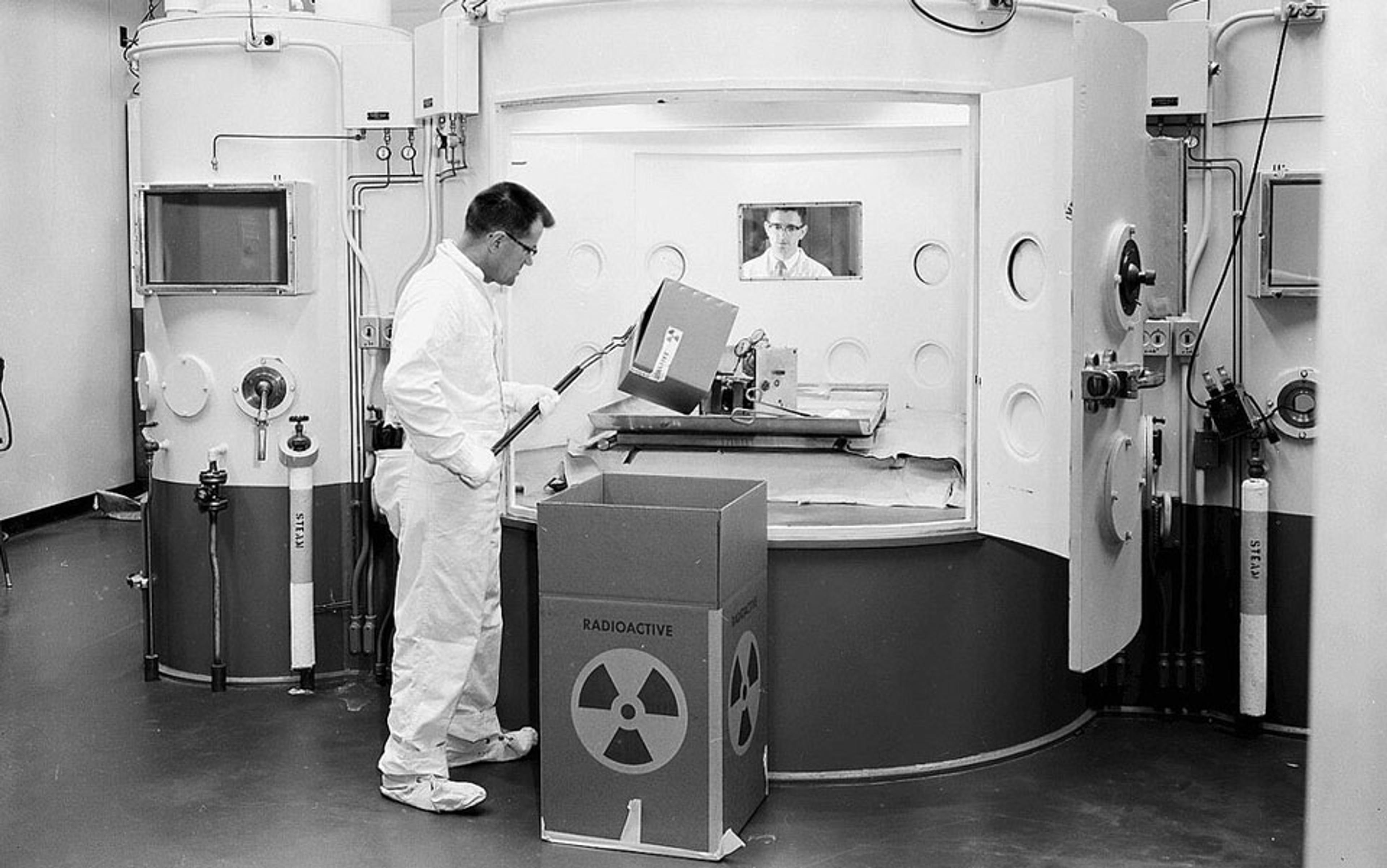 Photo of a man in a lab coat handling a radioactive box in a lab, standing by a large machine with a window and control panel.