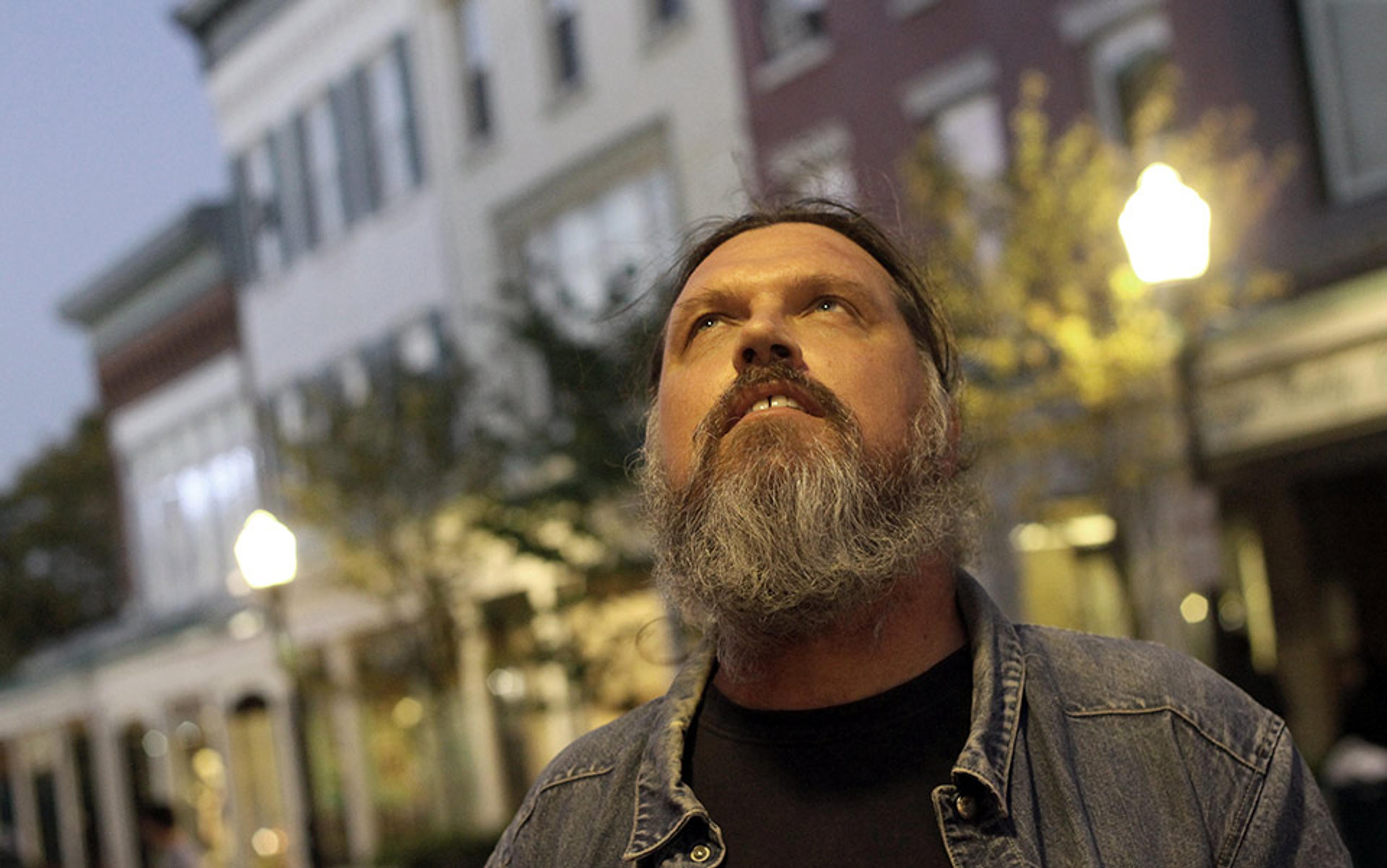 A bearded man in a denim jacket looking up, in front of an urban street with buildings and streetlights in the background.