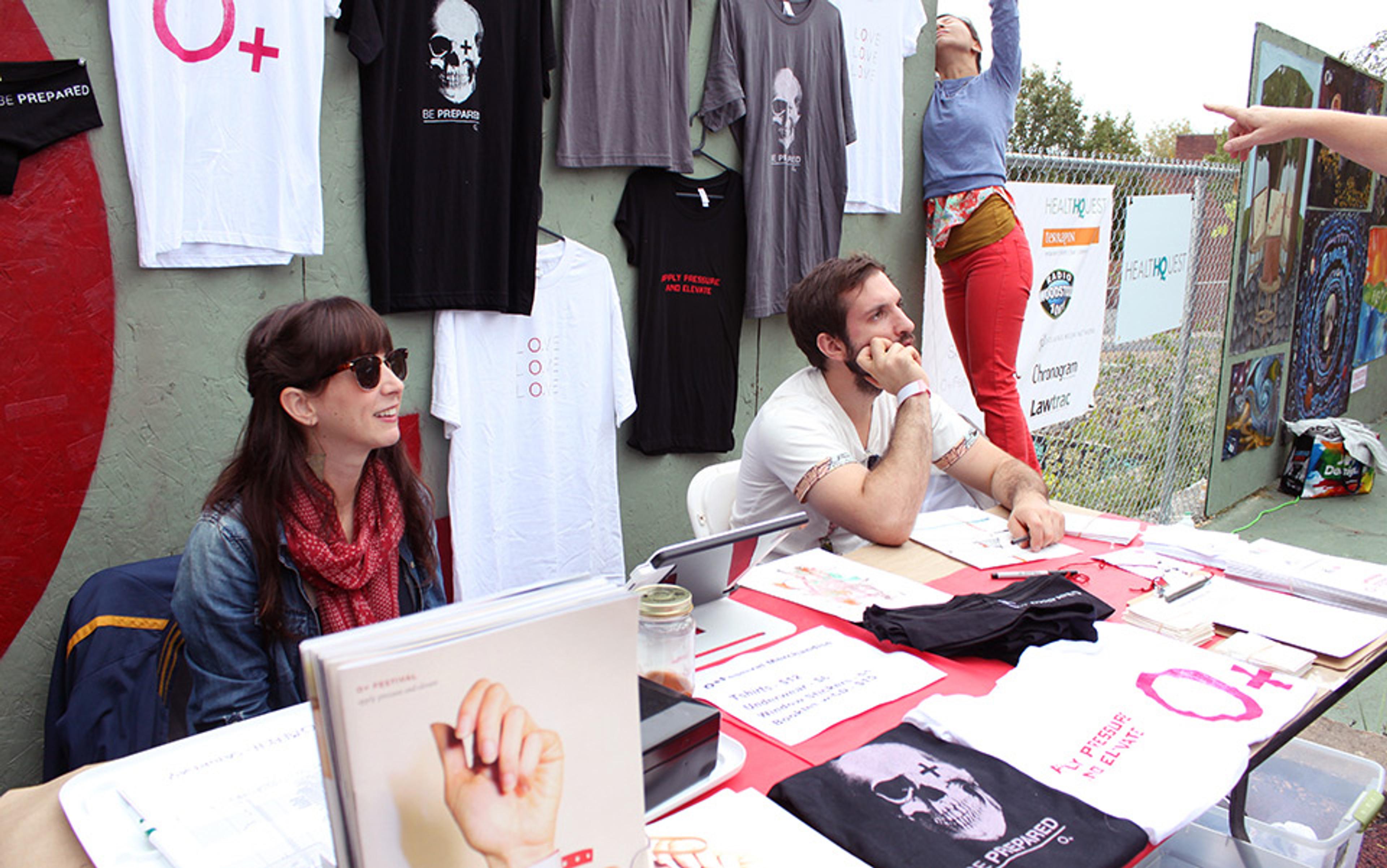 Two people at a stall with t-shirts on display. One person is sitting smiling, the other is resting their head on their hand.