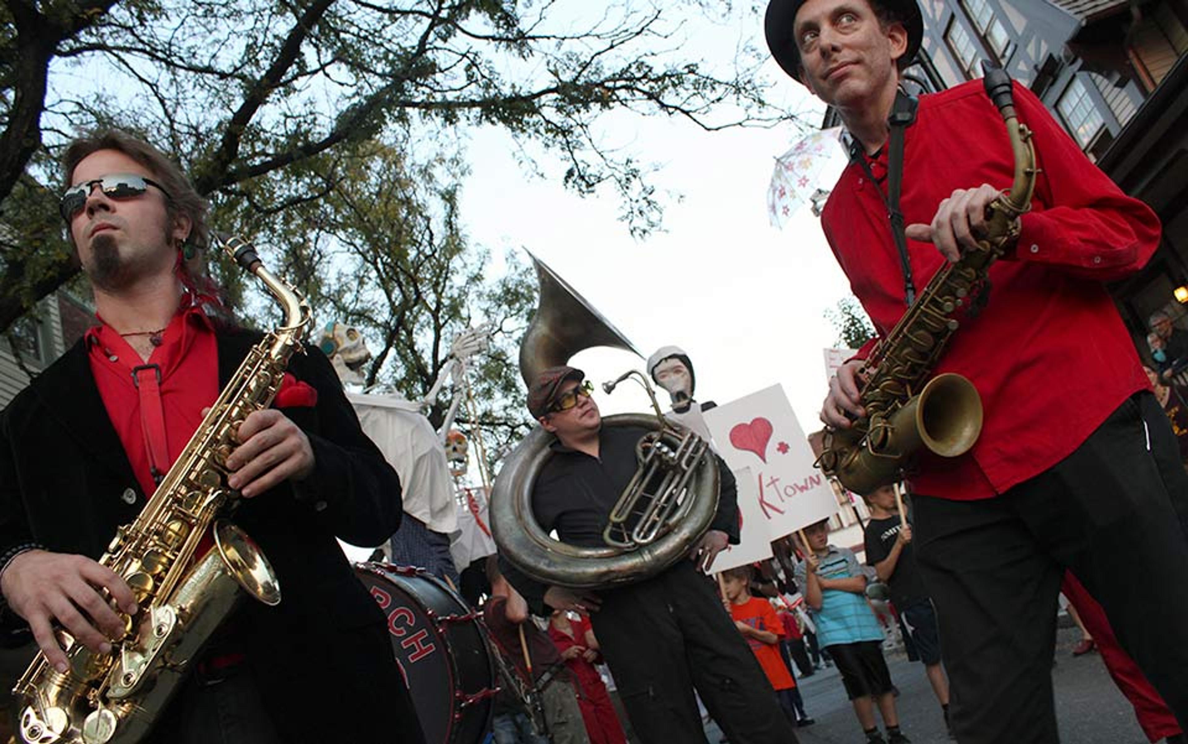 A street parade with musicians playing saxophones and a sousaphone, while people in the background hold signs and balloons.