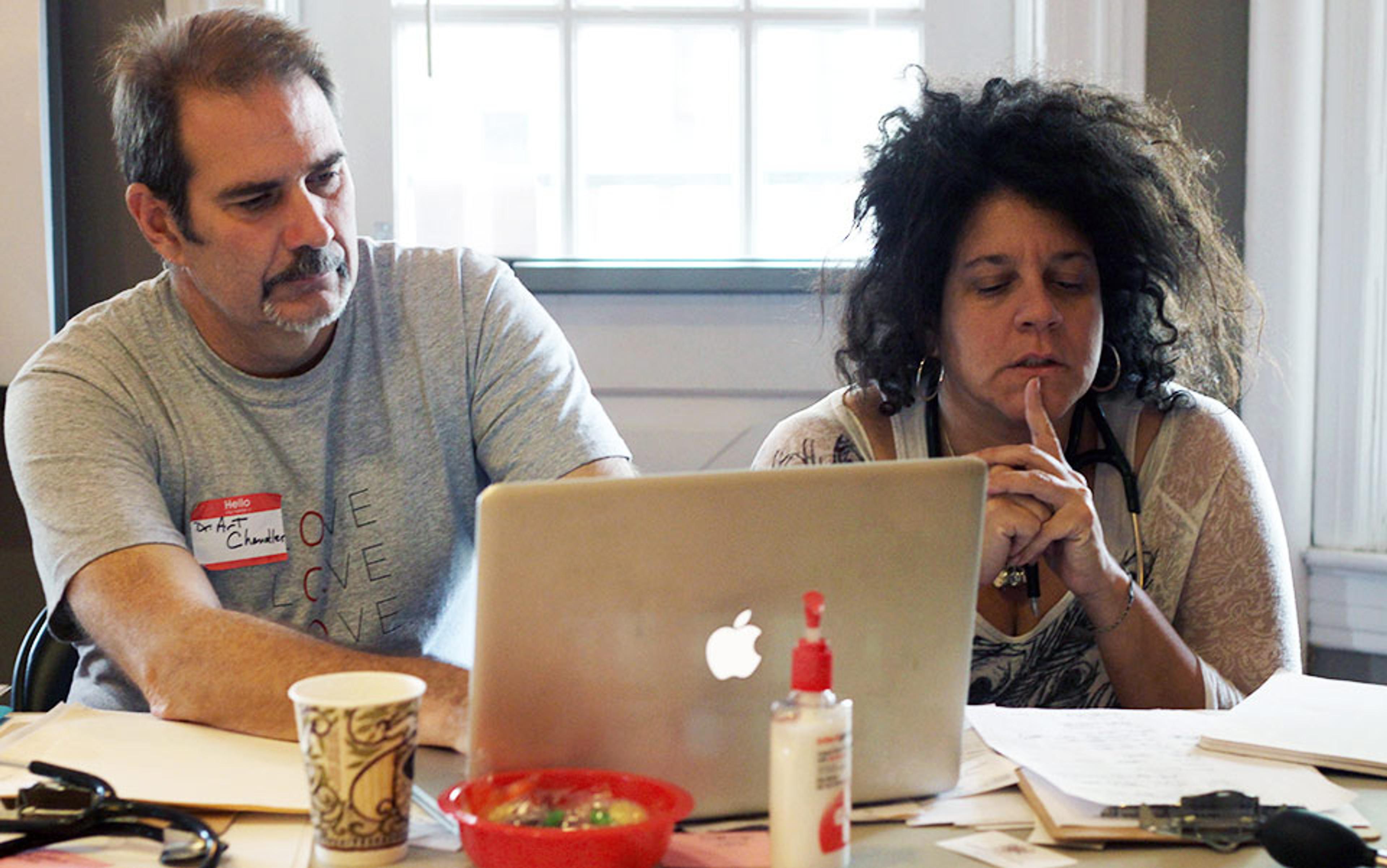 A man and woman sitting at a table using a laptop, with papers and a bowl in the foreground, in a bright room.