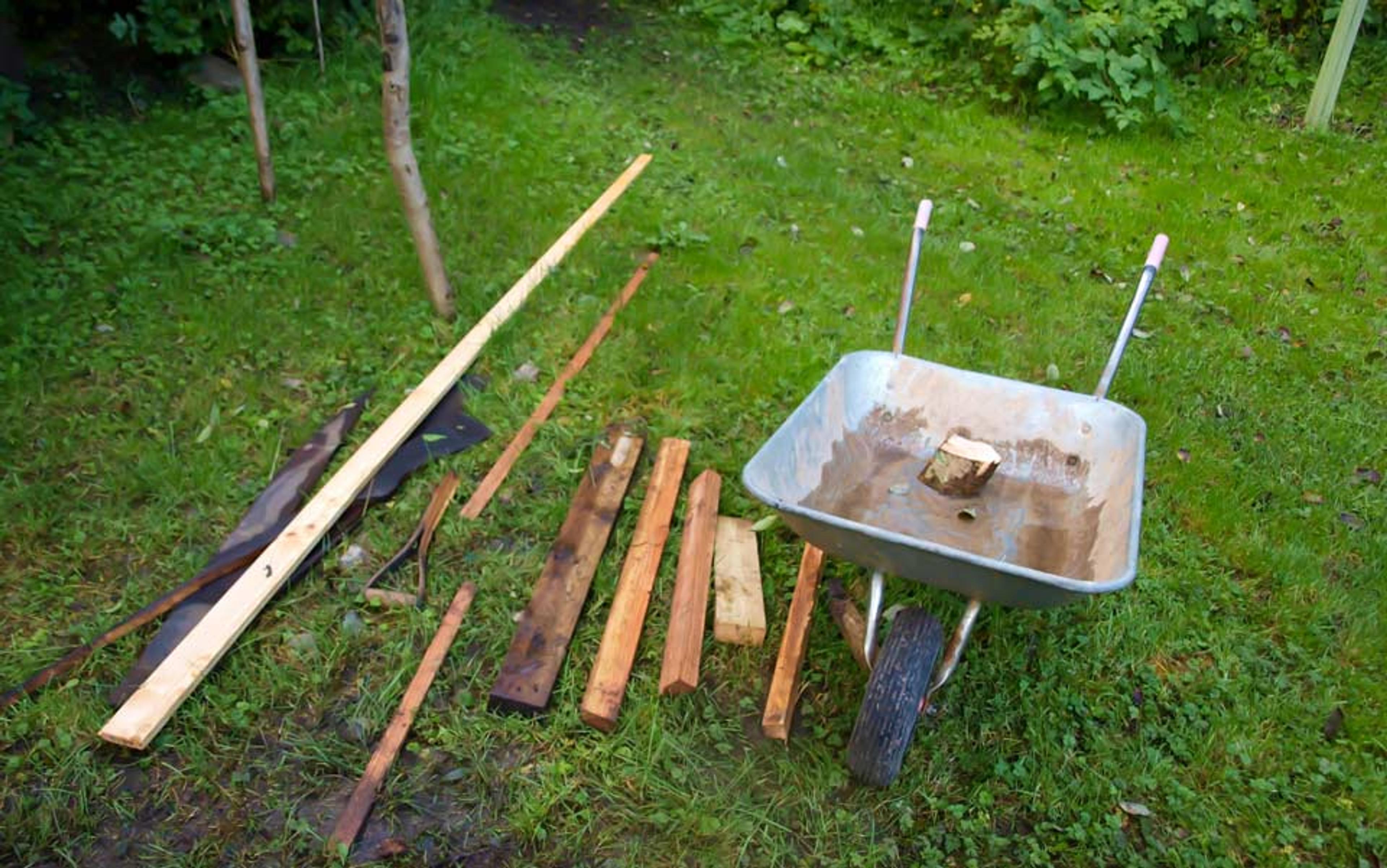 A wheelbarrow on grass next to various pieces of wood arranged on the ground in a garden setting.