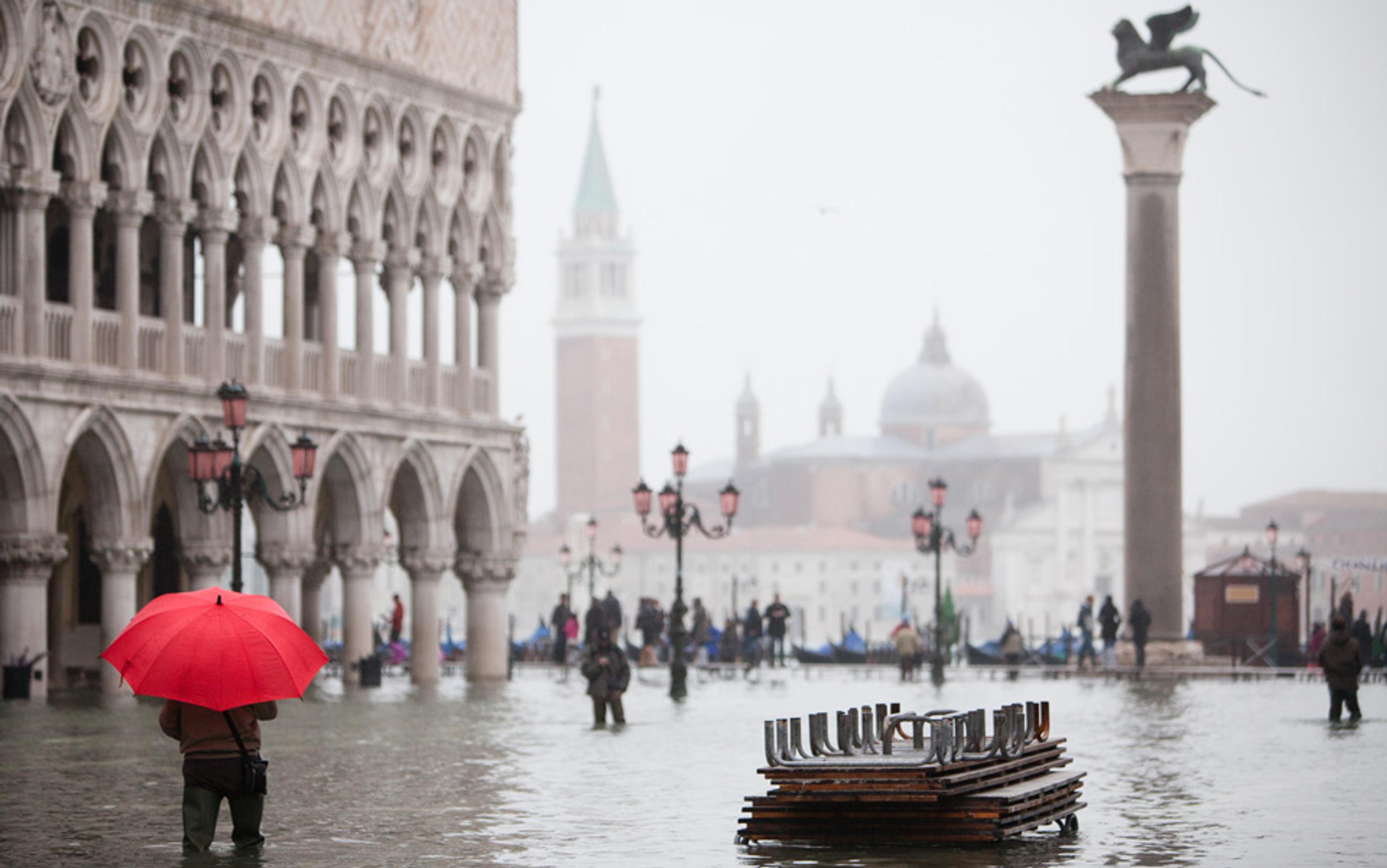 A flooded St Mark’s Square in Venice. A person with a red umbrella stands up to his knees in water near a historic building and a tall column with a statue.