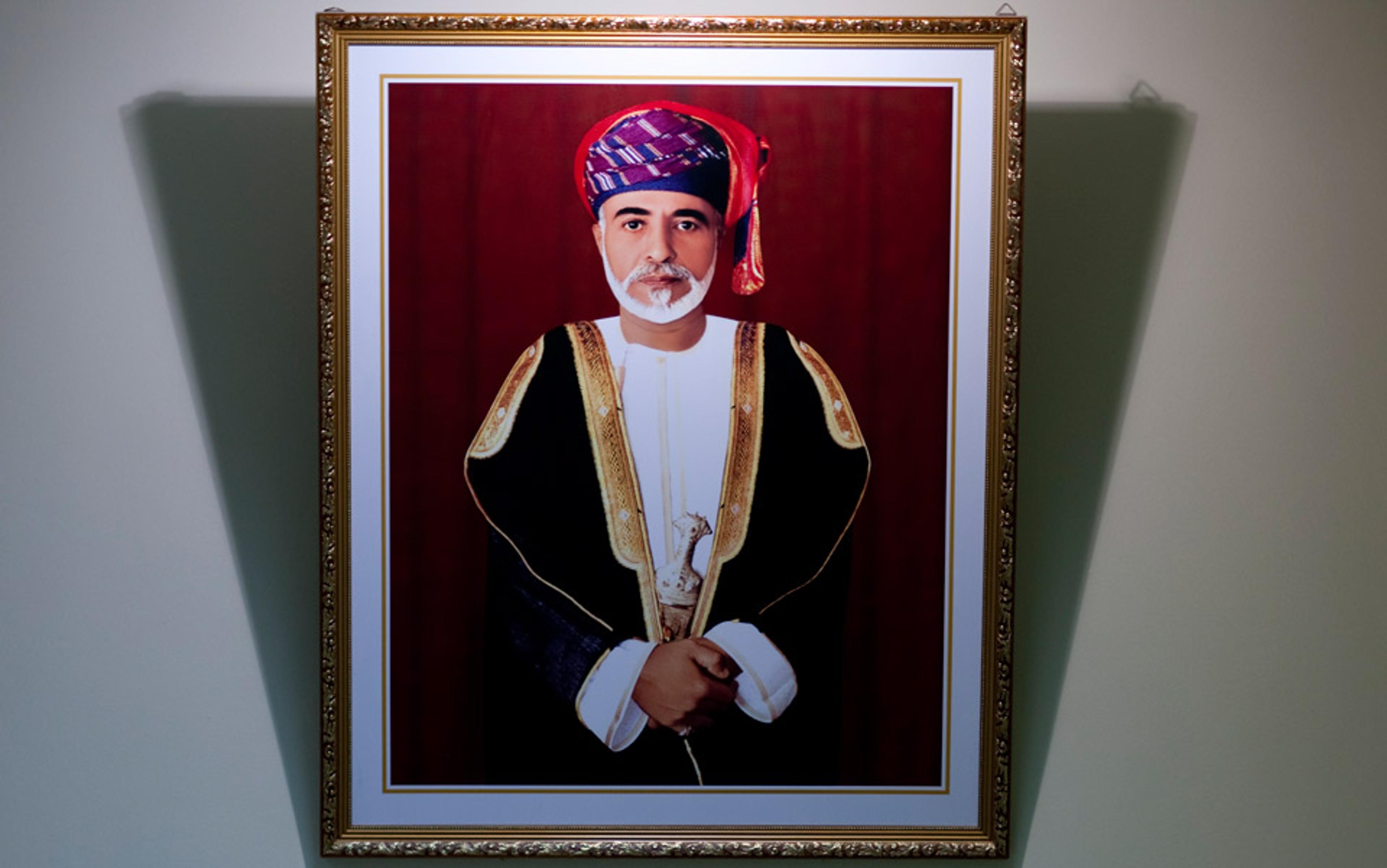 Framed photo of a man in traditional attire with a red background, traditional cap and black robe, hands clasped, hung on a wall.