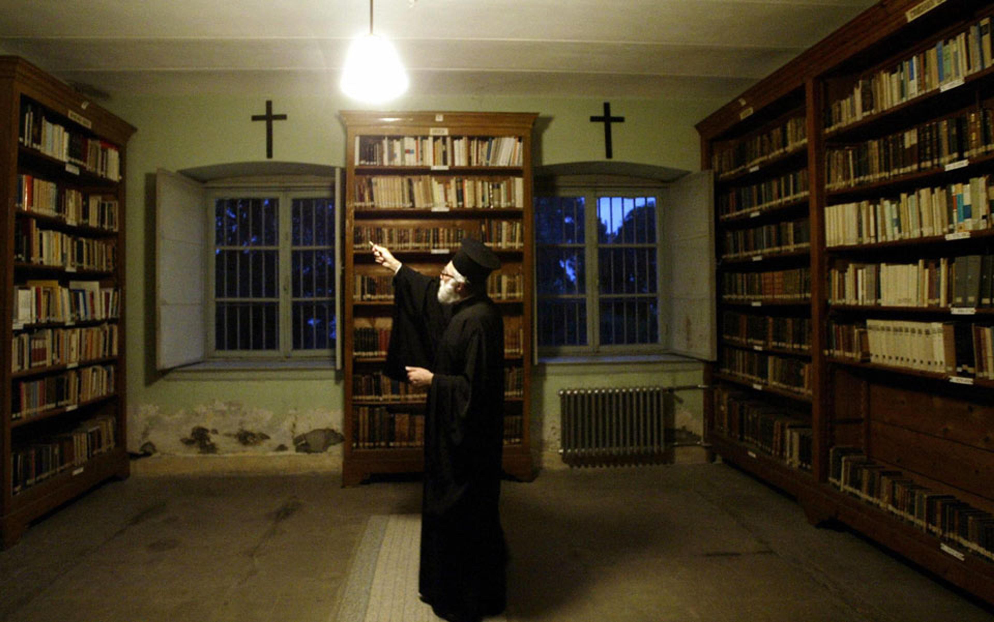 Photo of a man in a long robe pointing at a bookshelf in a dimly lit library with crosses above the windows and shelves.