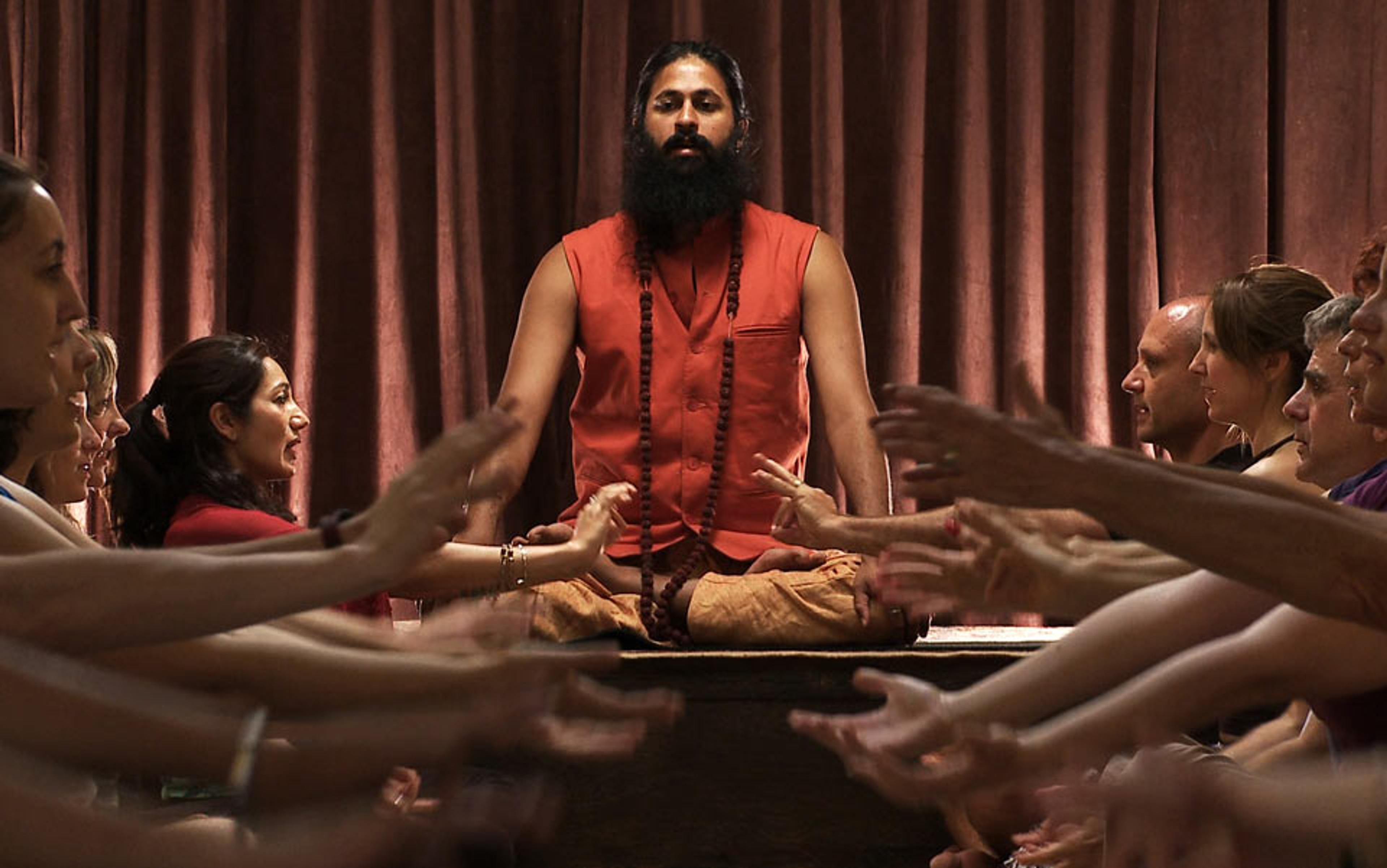 A bearded man in orange robes meditating on a platform with arms raised, surrounded by people in a class environment.