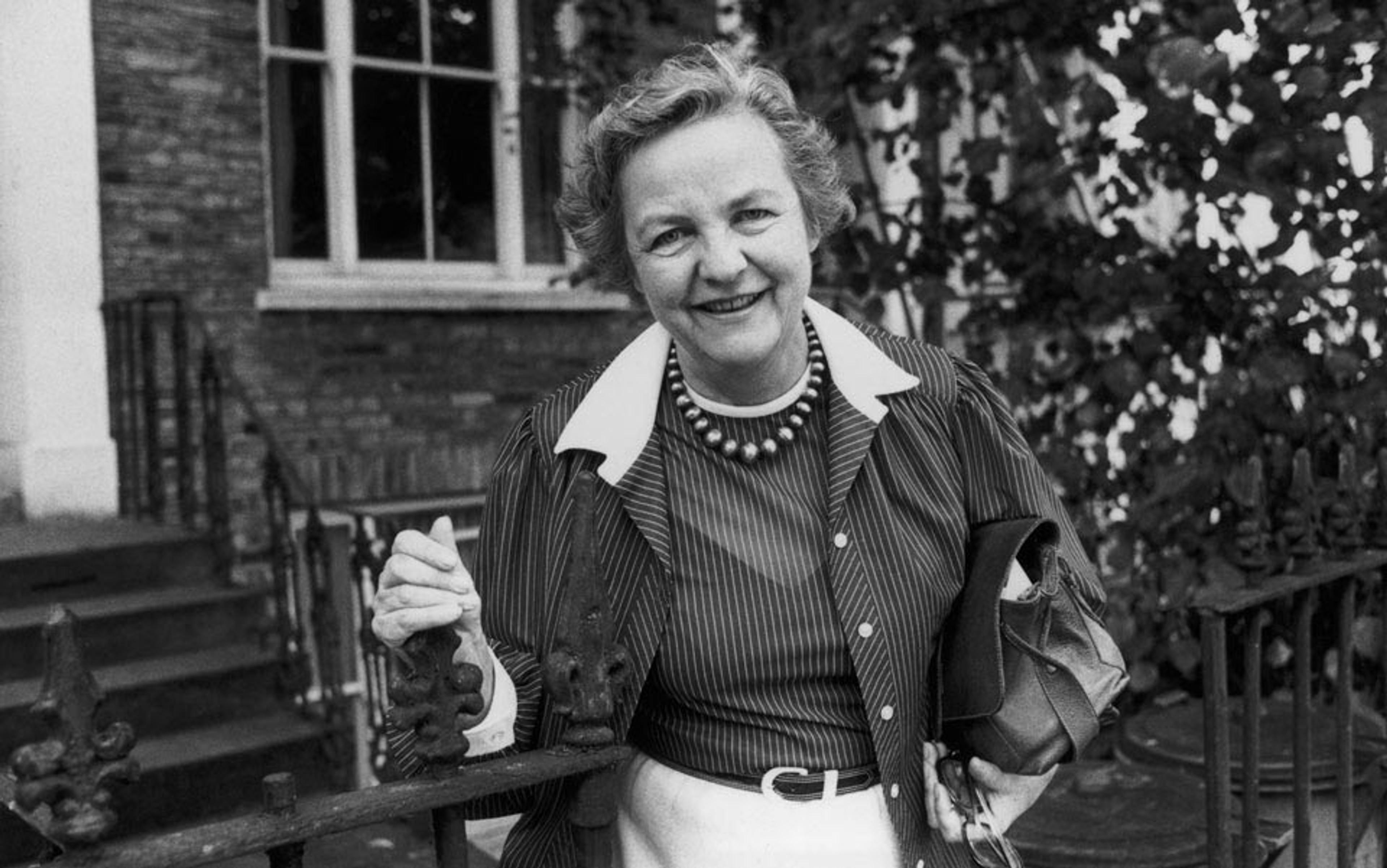 Black and white photo of a smiling woman holding a gate in front of a brick house with a window, wearing a striped blouse and pearl necklace.