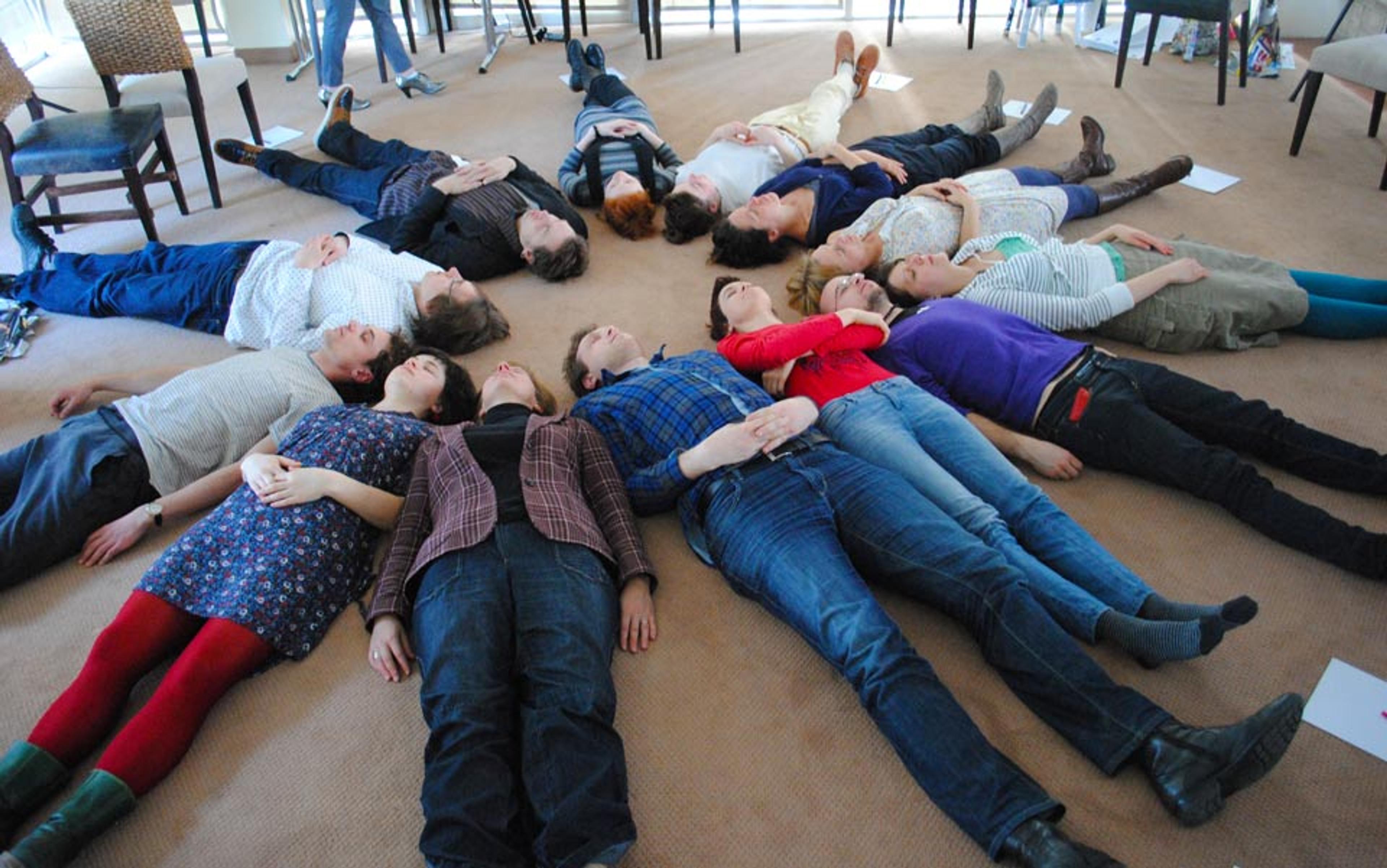 A group of people lying on their backs in a circle on a carpeted floor with their heads pointed towards the centre.