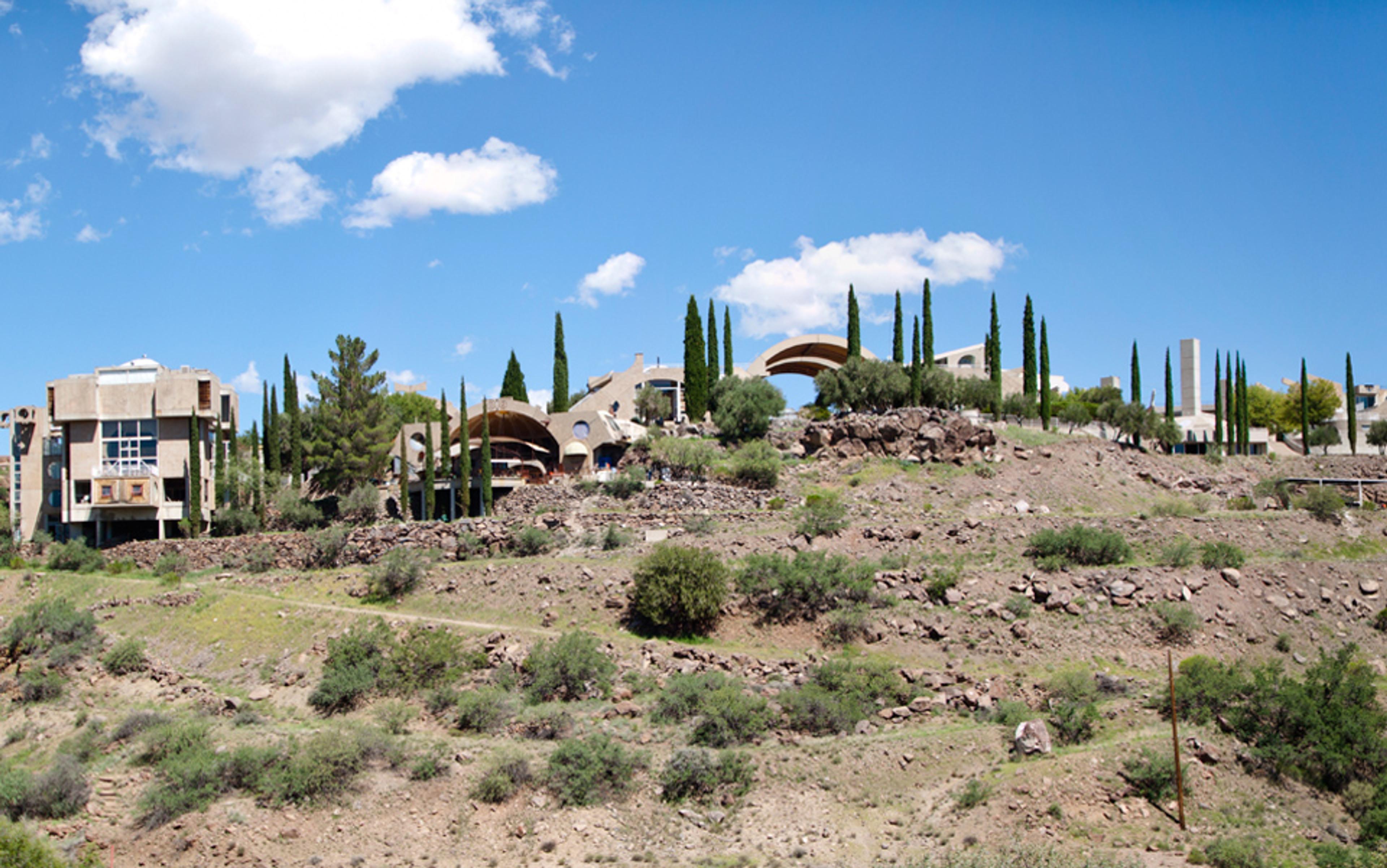 Photo of a hillside with unique modern buildings and tall cypress trees against a clear blue sky with scattered clouds.