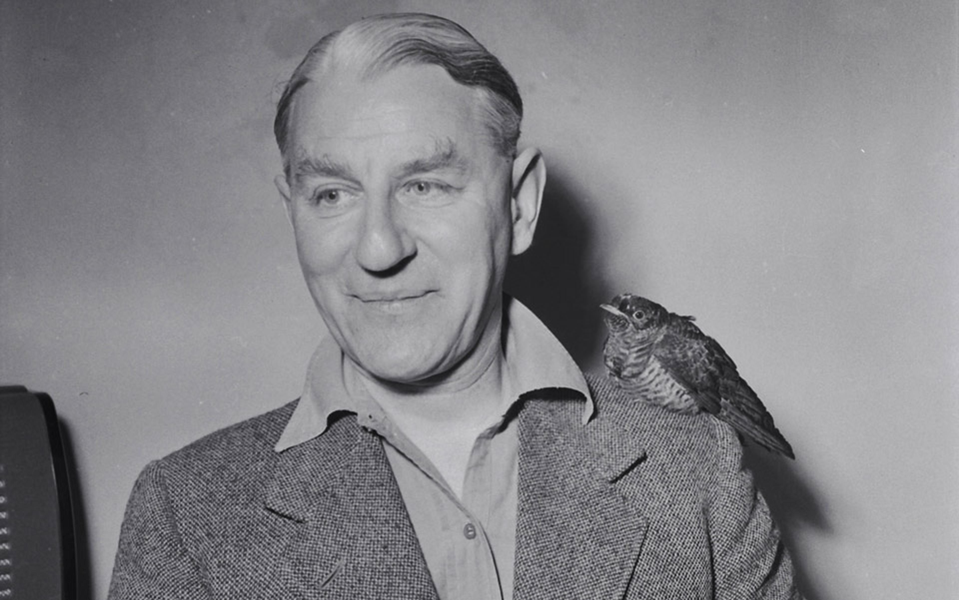 Black and white photo of a man smiling with a bird perched on his shoulder; the background is plain and the man wears a suit jacket.