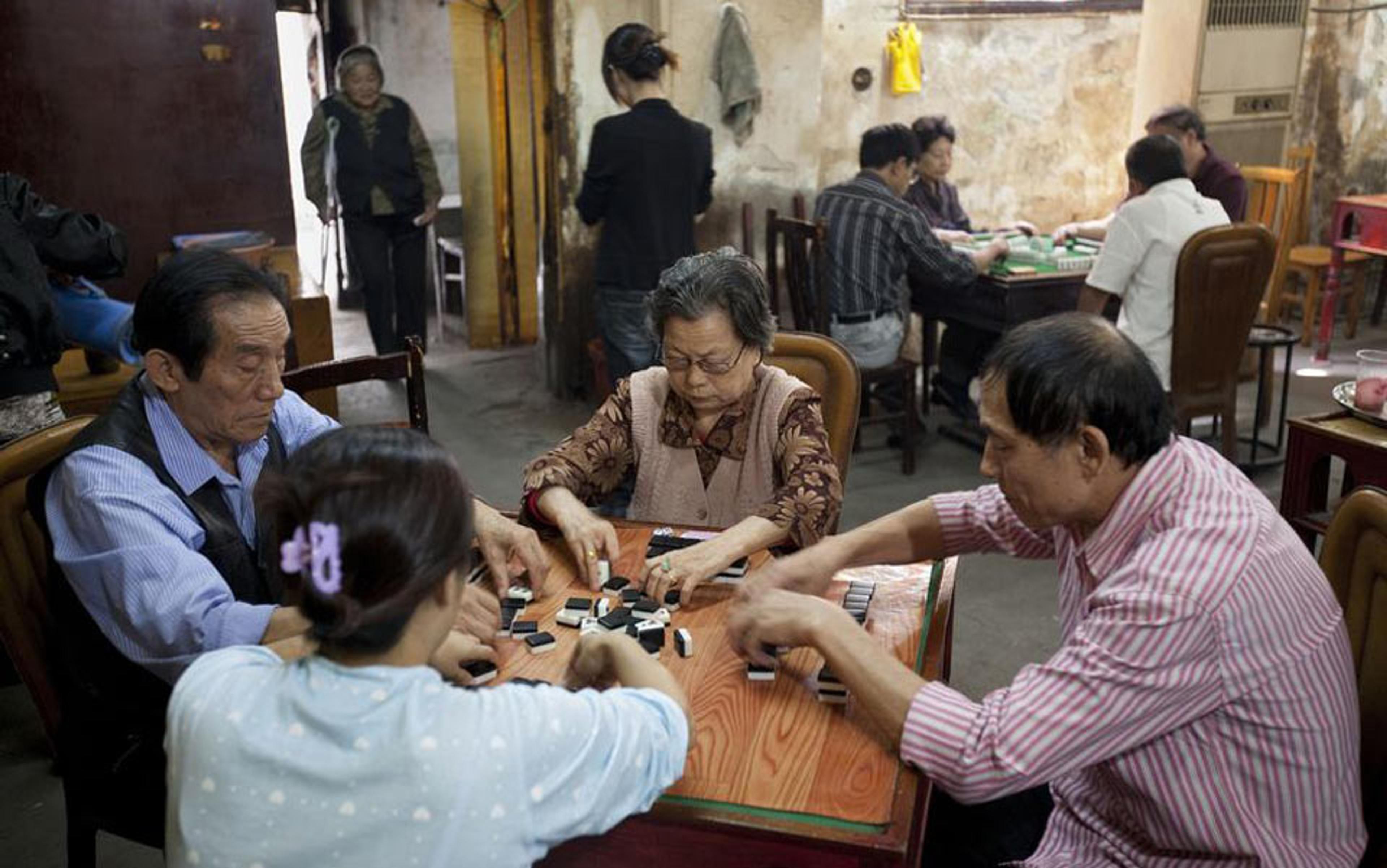 Elderly Chinese people playing mahjong at a table in a room with more people playing in the background.