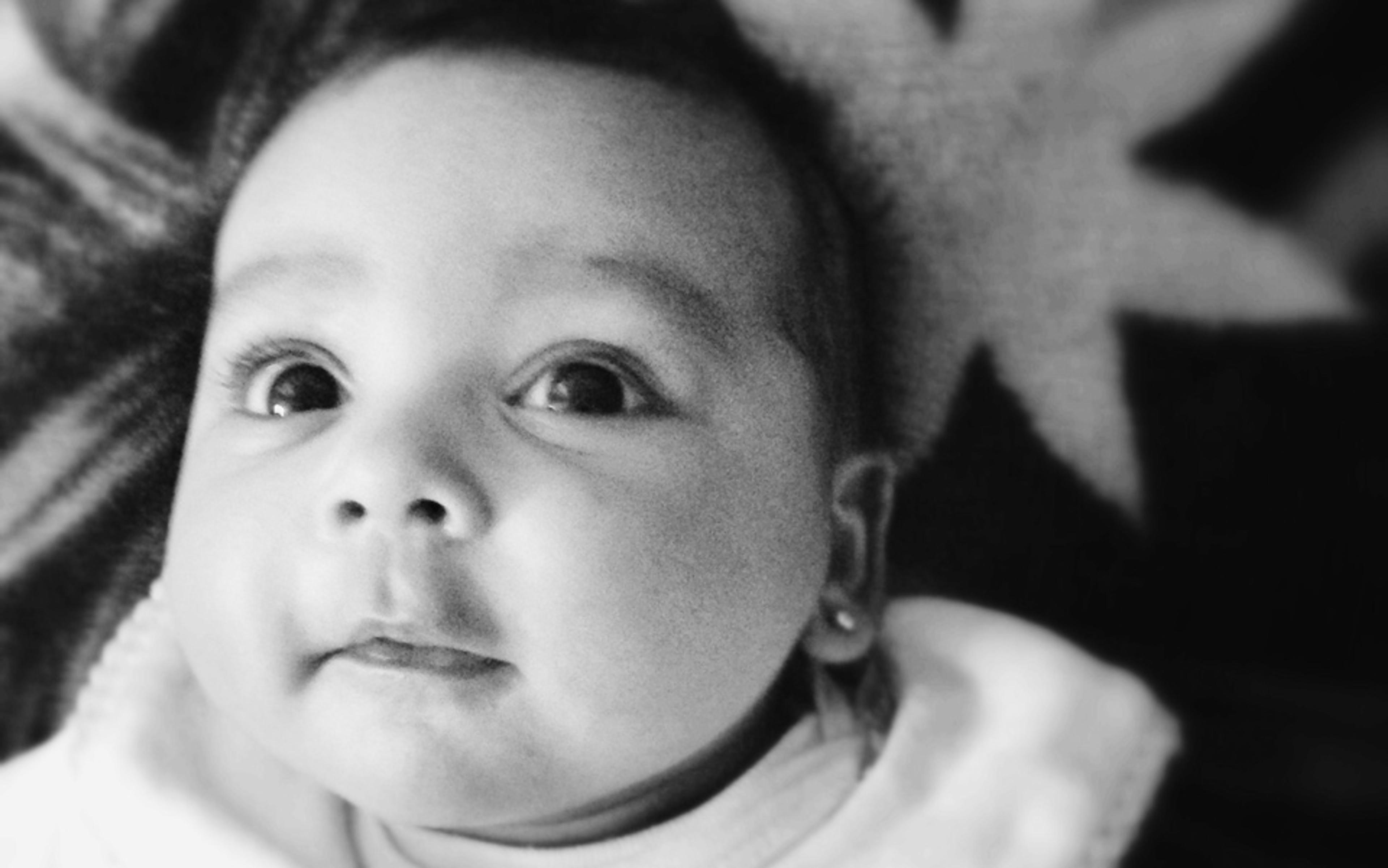 Black and white photo of a baby lying down, looking at the camera with wide eyes and a calm expression, wearing a white outfit.