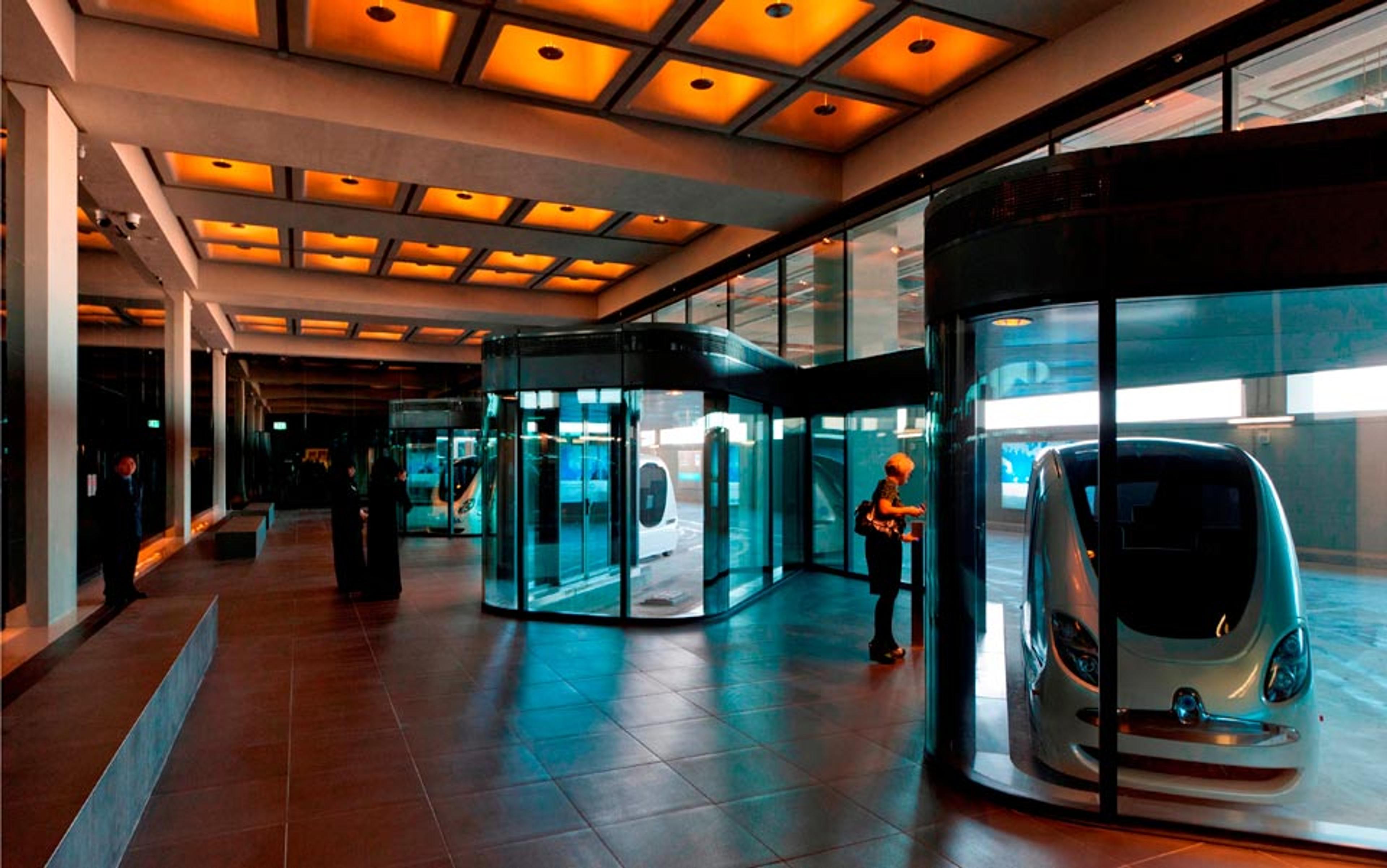 Photo of a modern indoor space with glass walls displaying futuristic vehicles, illuminated by warm ceiling lights, with people observing.