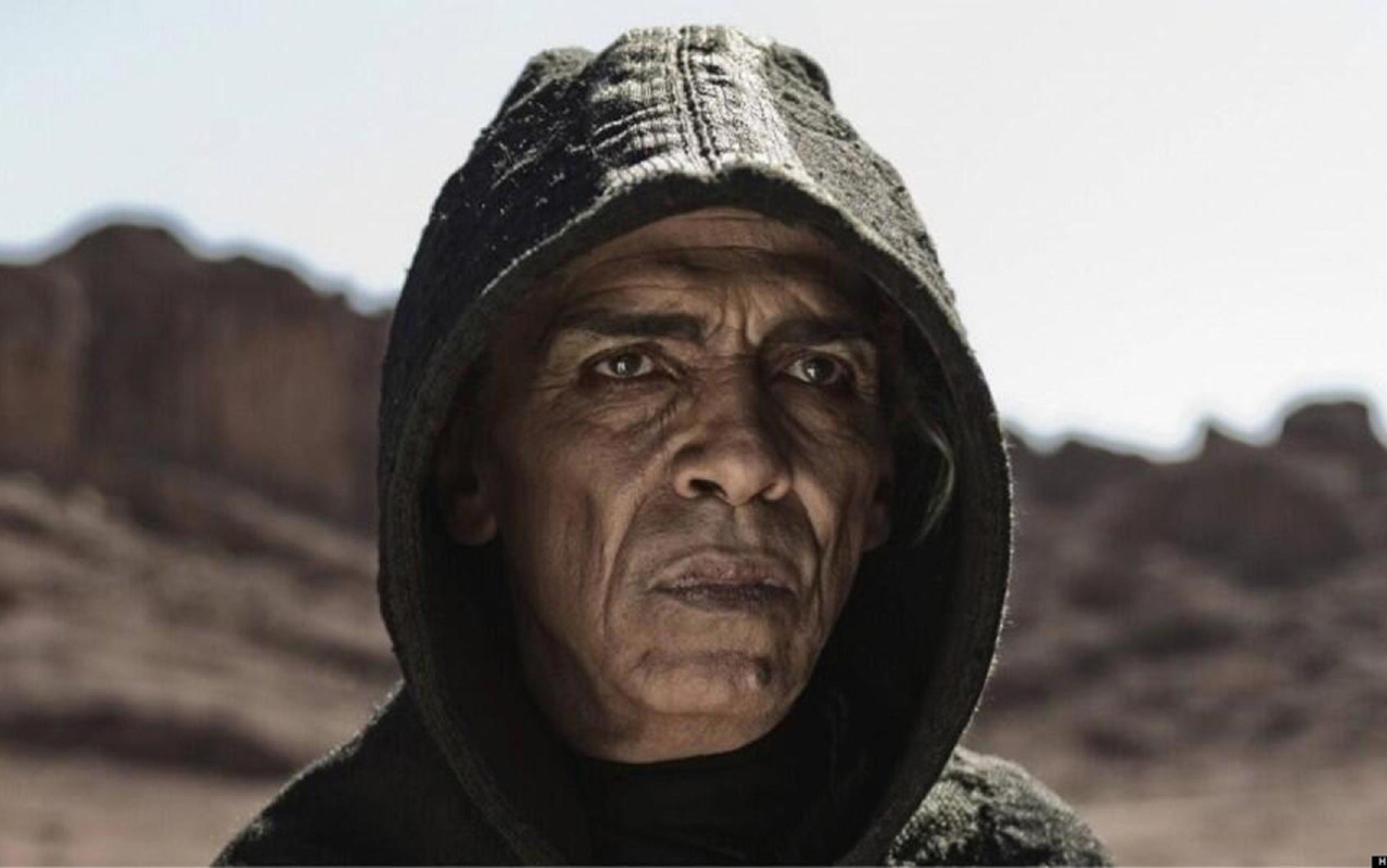 Photo of a man in a dark hooded cloak, with a serious expression, standing outdoors with rocky desert landscape in the background.