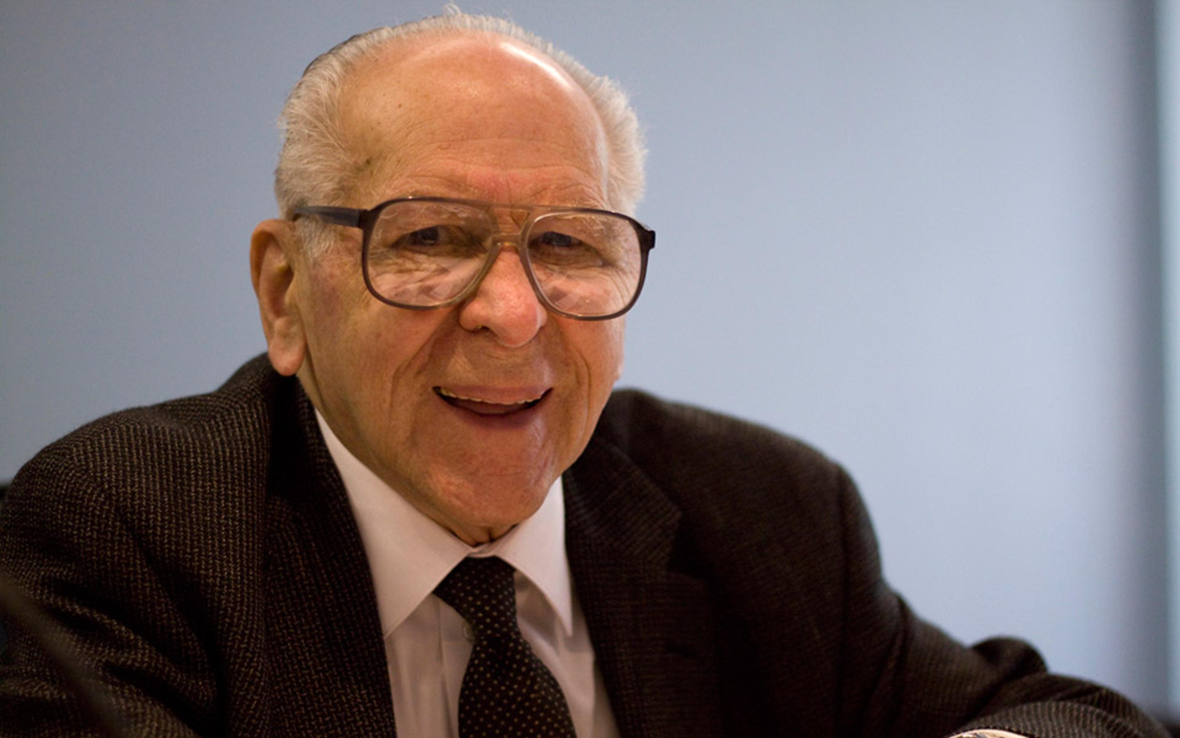 An elderly man wearing glasses and a suit, smiling, against a plain background. The lighting is soft, creating a warm atmosphere.