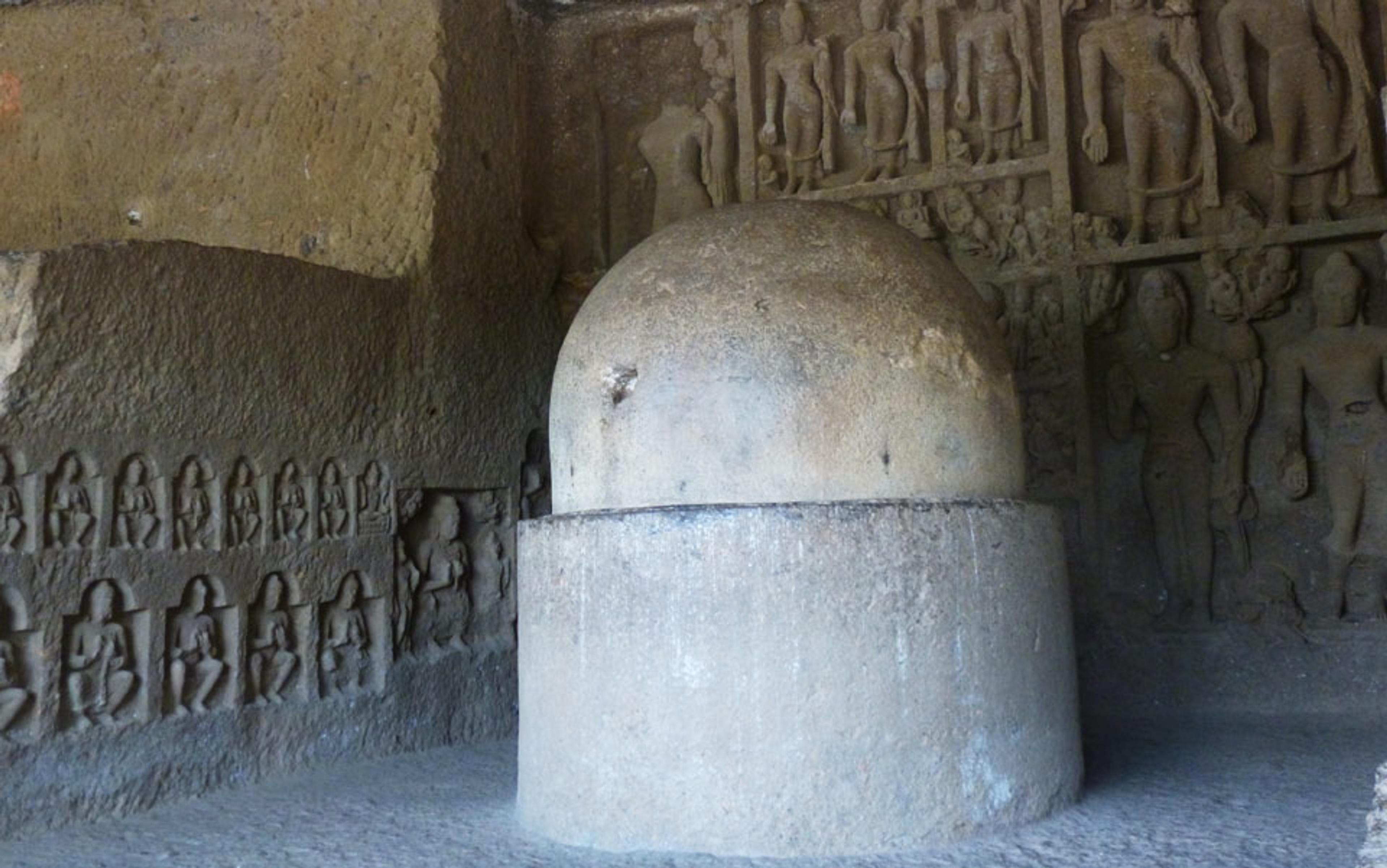 Photo of an ancient stone stupa inside a cave with intricate wall carvings depicting figures and deities in relief.