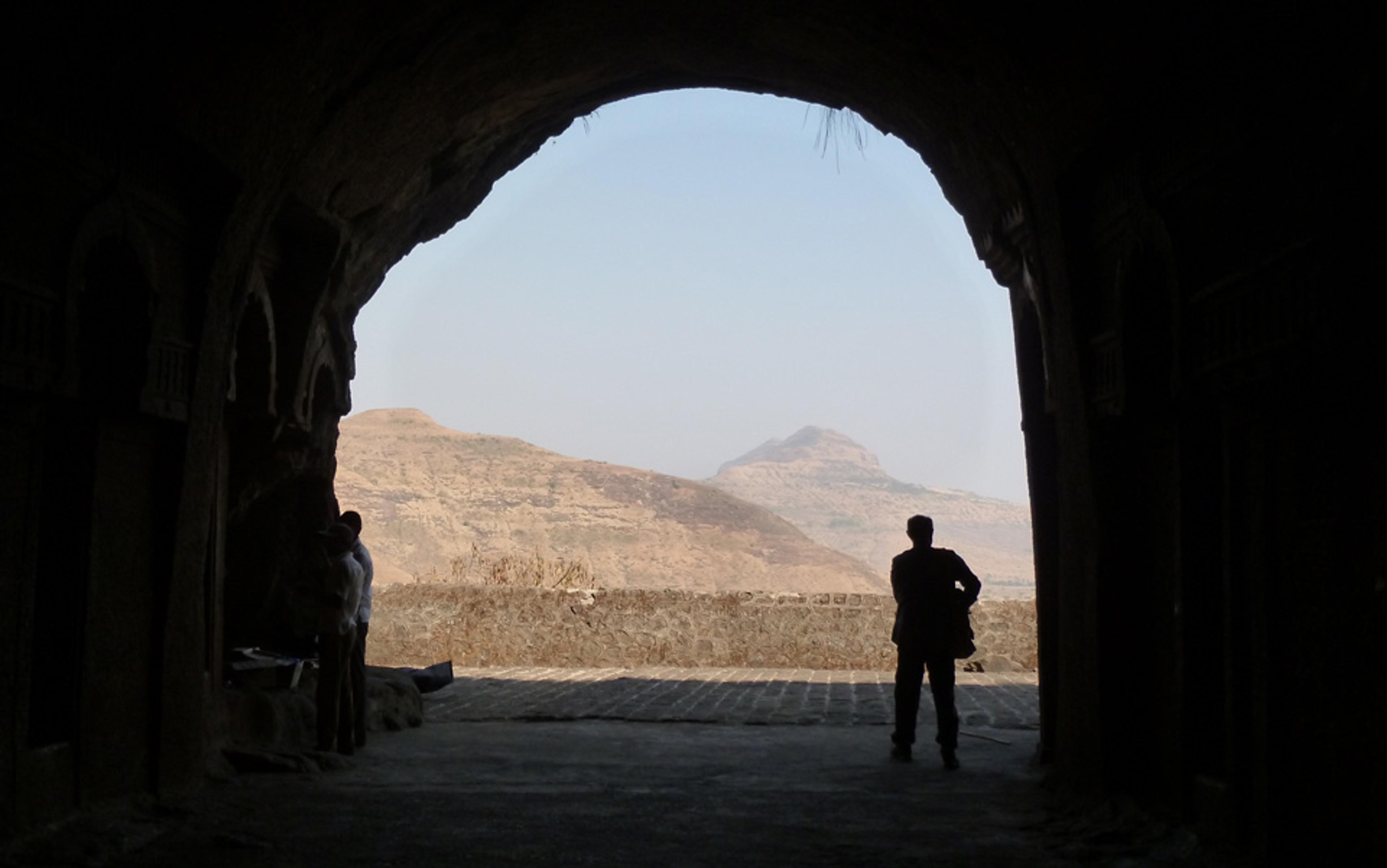 Photo of a silhouetted person standing in a cave entrance, looking out at a mountainous landscape under a clear sky.