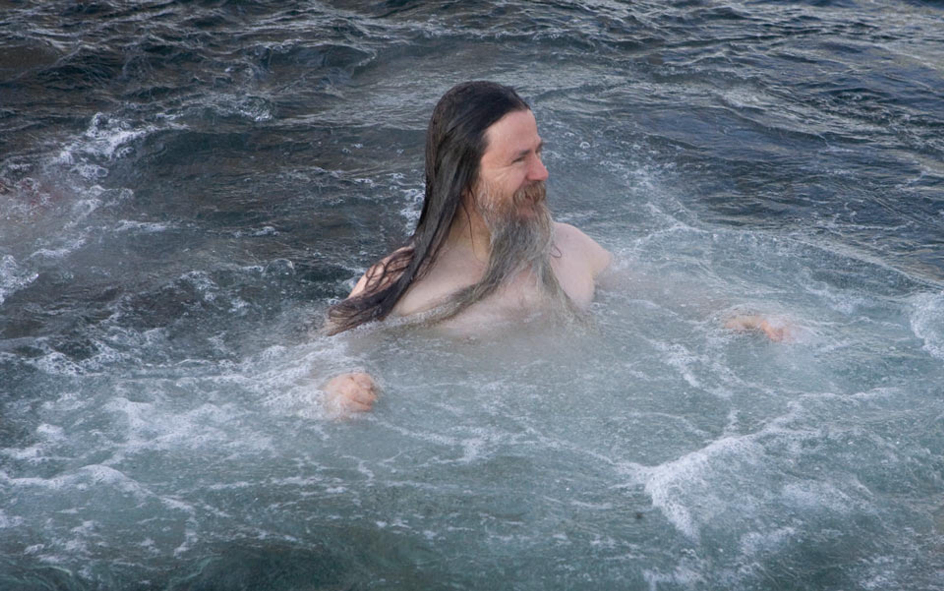 A photo of a middle-aged man with long hair and a beard swimming in a body of water, with waves and splashes around him.