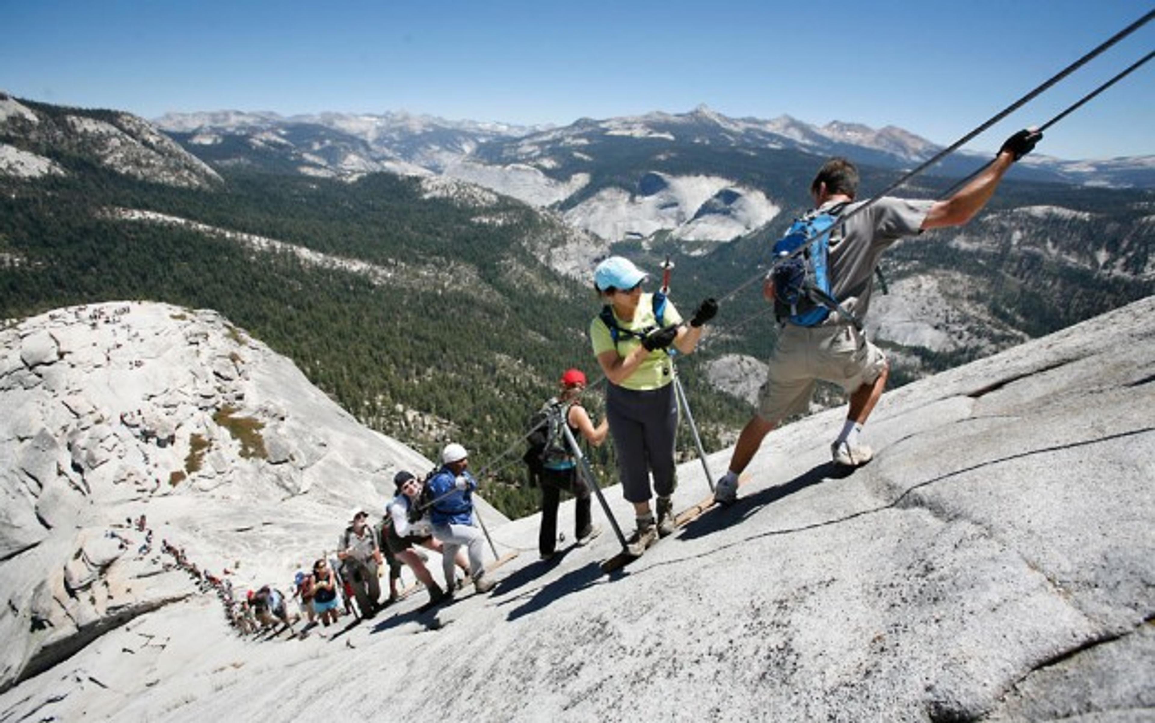 Hikers using cables to ascend a steep rock face with a mountainous landscape in the background under a clear blue sky.