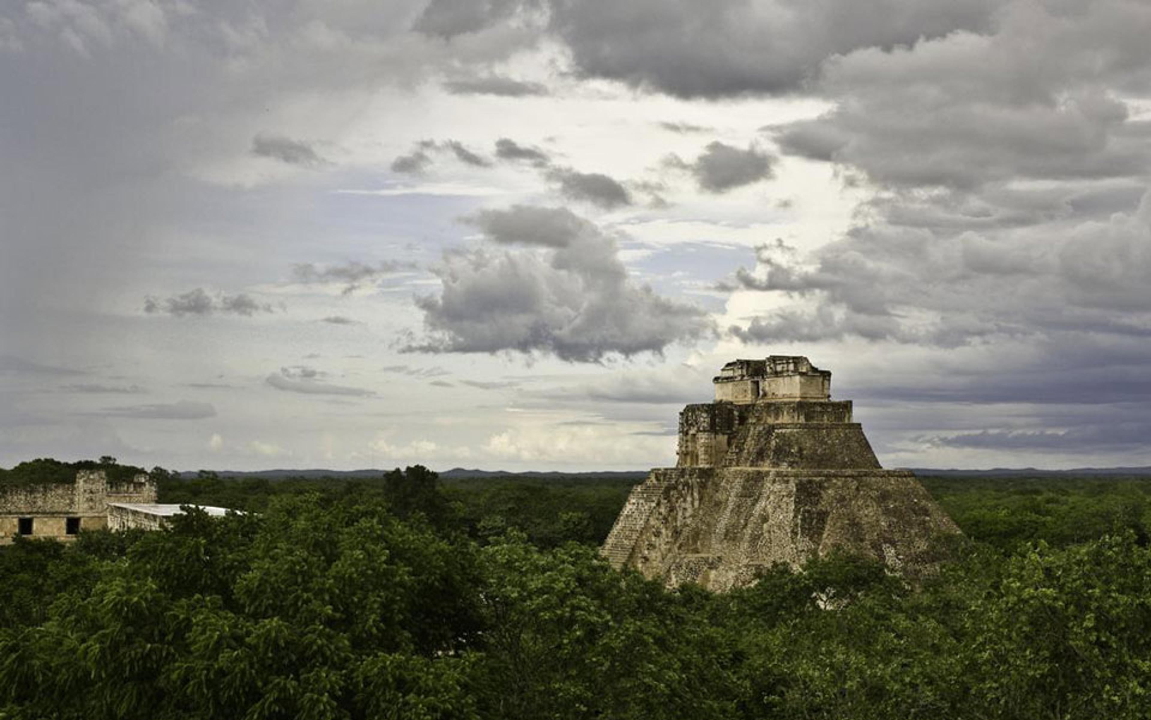 An ancient stone pyramid surrounded by green forest under a cloudy sky, showcasing ruins in the foreground.