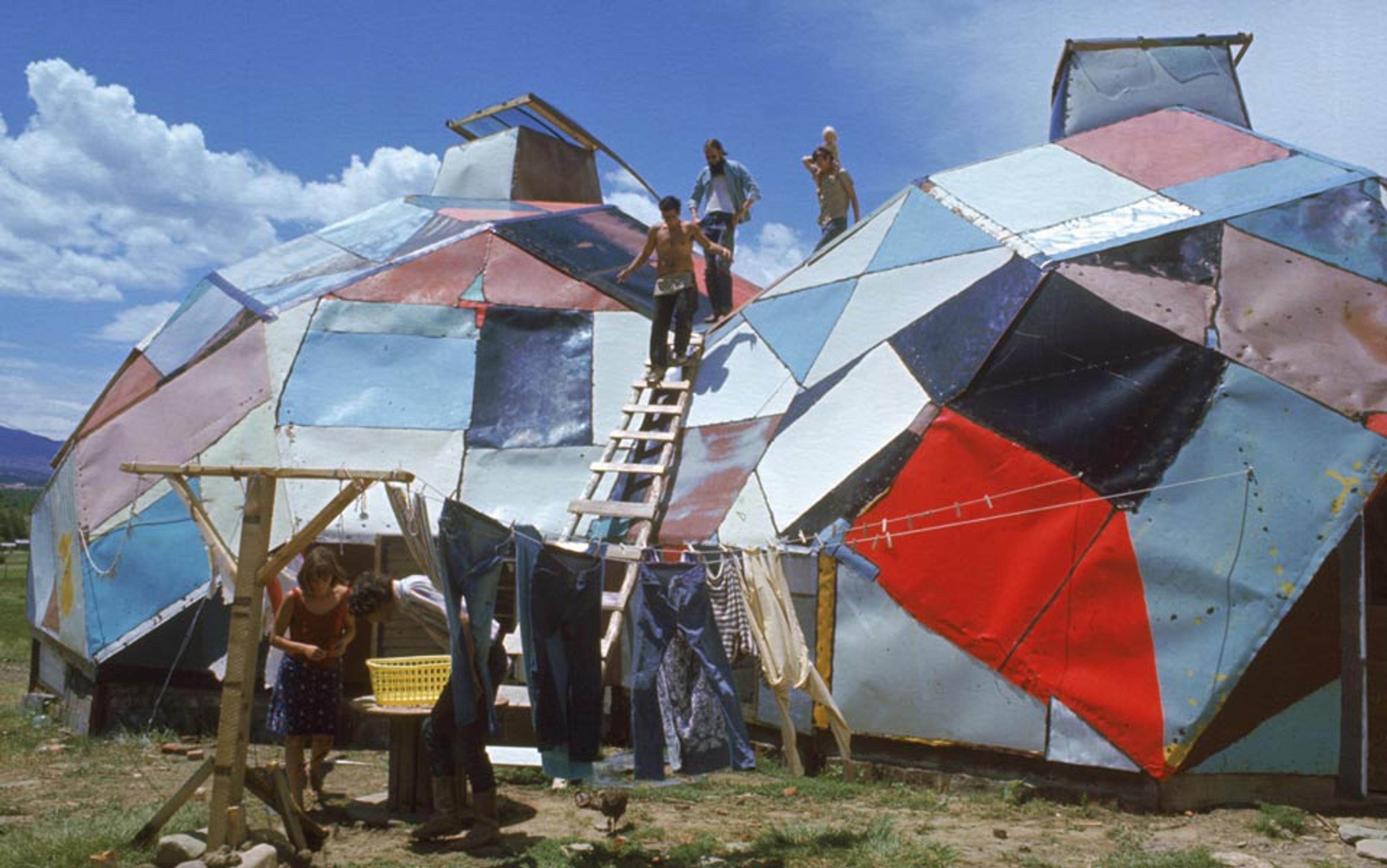 Photo of several people outside a colourful, geometrically patched dome. Some are on the roof, others tend to laundry drying on a line.