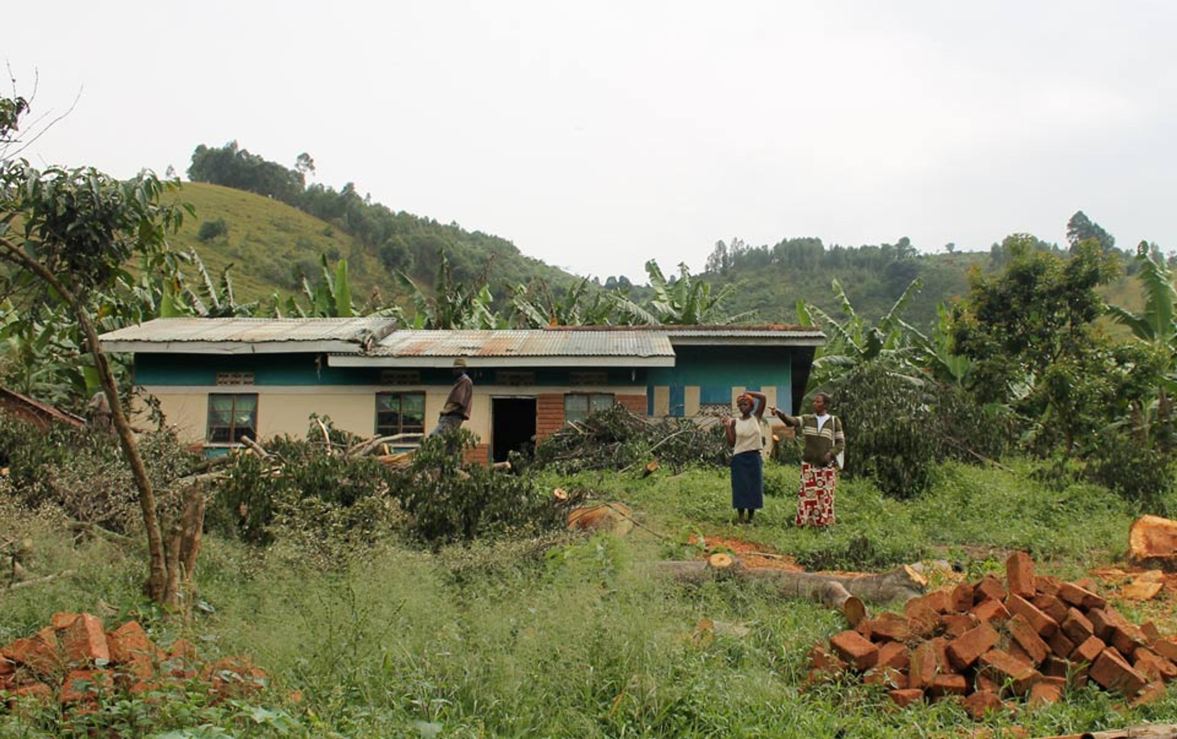 A rural house with a tin roof in a hilly area, surrounded by lush greenery. Three people stand in the front yard.