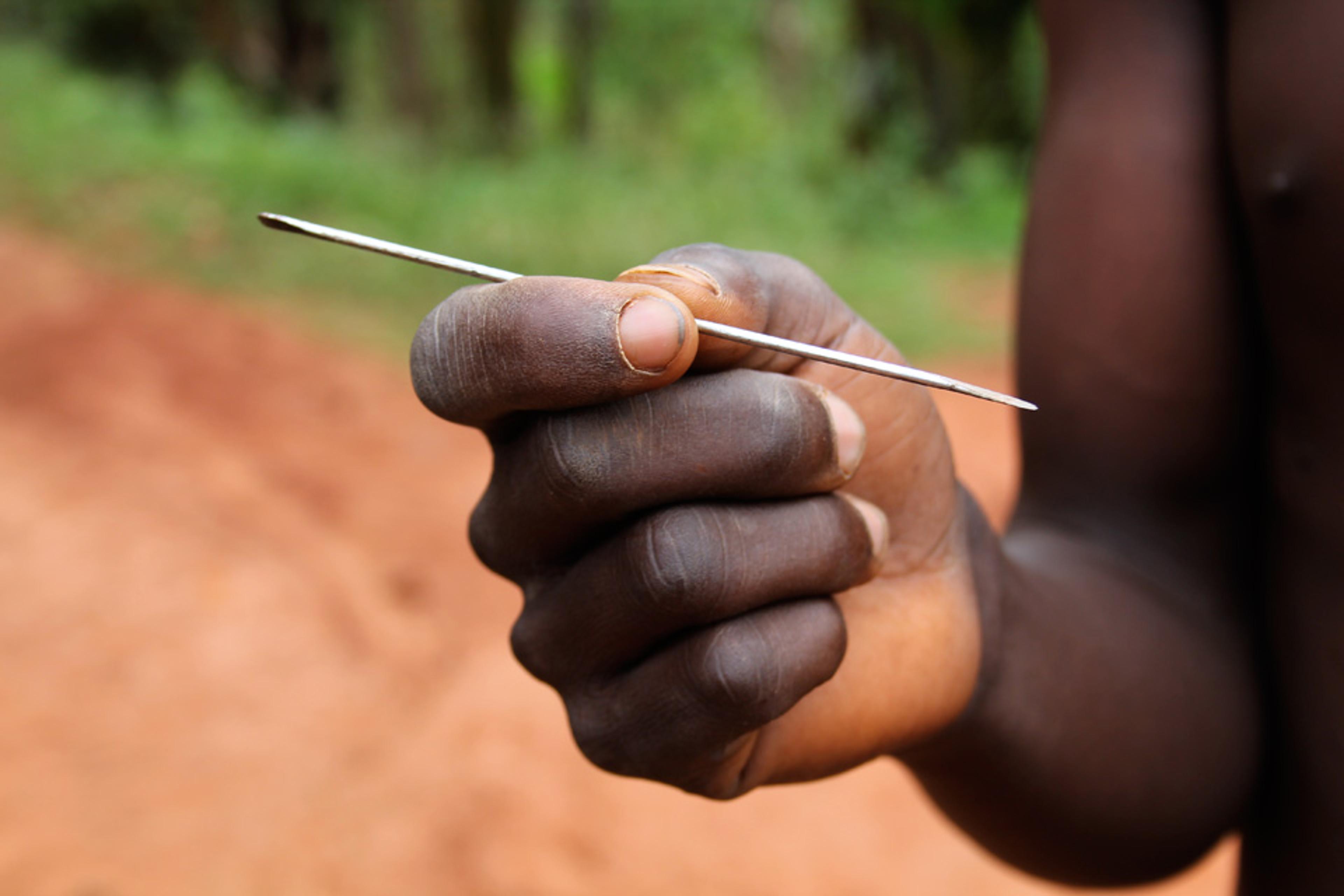 A close-up of a hand holding a thin metal tool with a blurred outdoor background featuring soil and greenery.