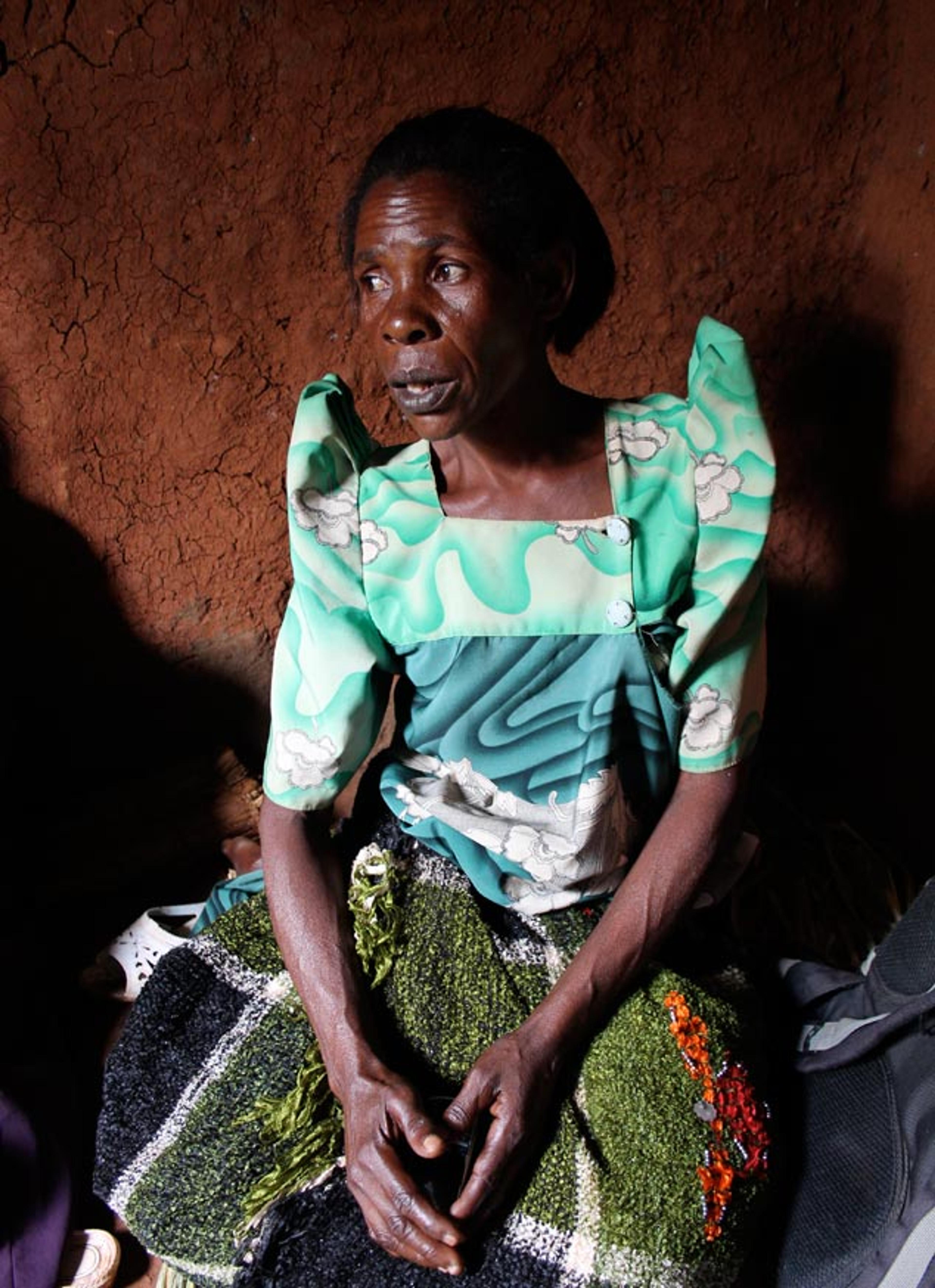 A Black woman in colourful clothes sitting against a textured, earth wall with hands clasped and looking to the side.