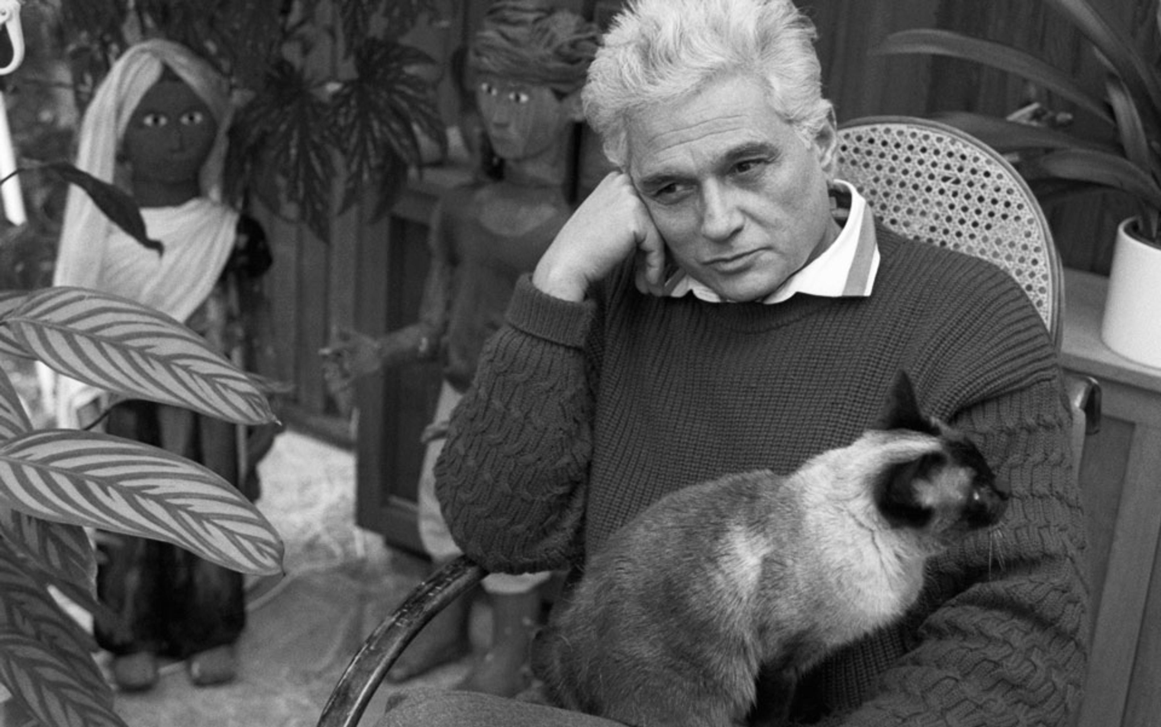 Black and white photo of a man with grey hair sitting with a cat on his lap, surrounded by plants and two decorative figurines.