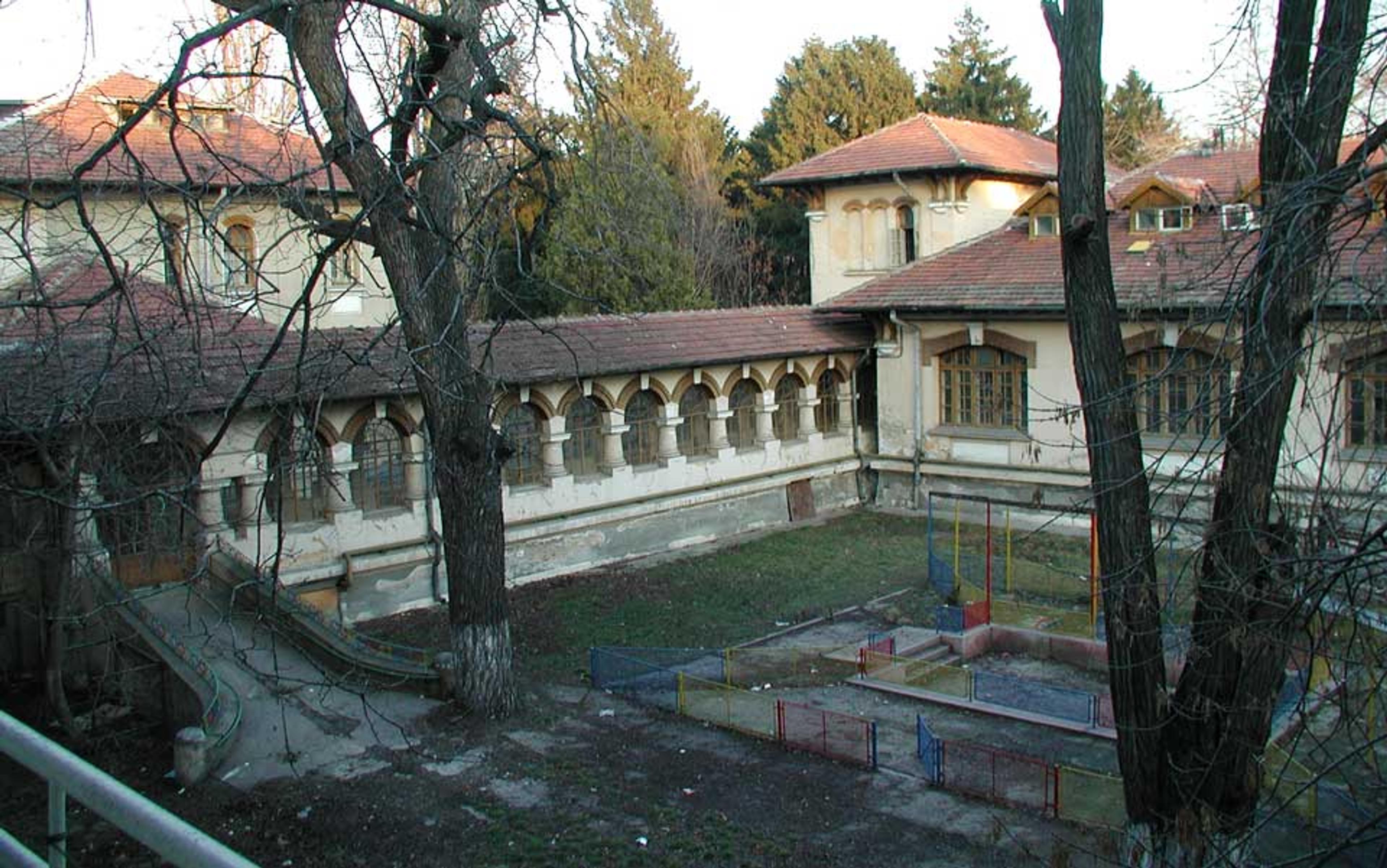 An old courtyard with arched windows, tiled roofs, bare trees, and a fenced area, surrounded by lush green trees in the background.