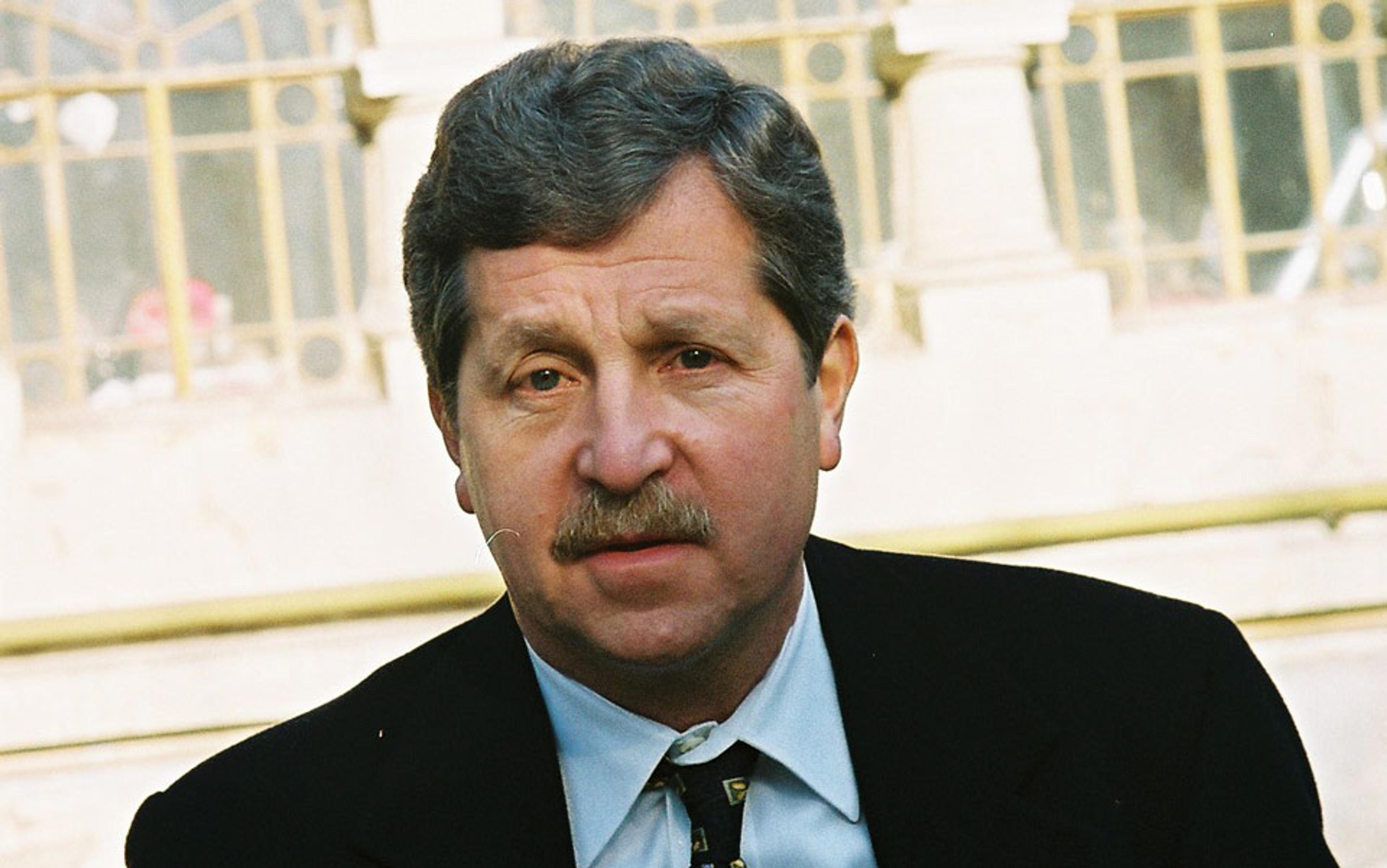 A man with greying hair and a moustache wearing a black suit and light blue shirt, standing in front of a building with columns.