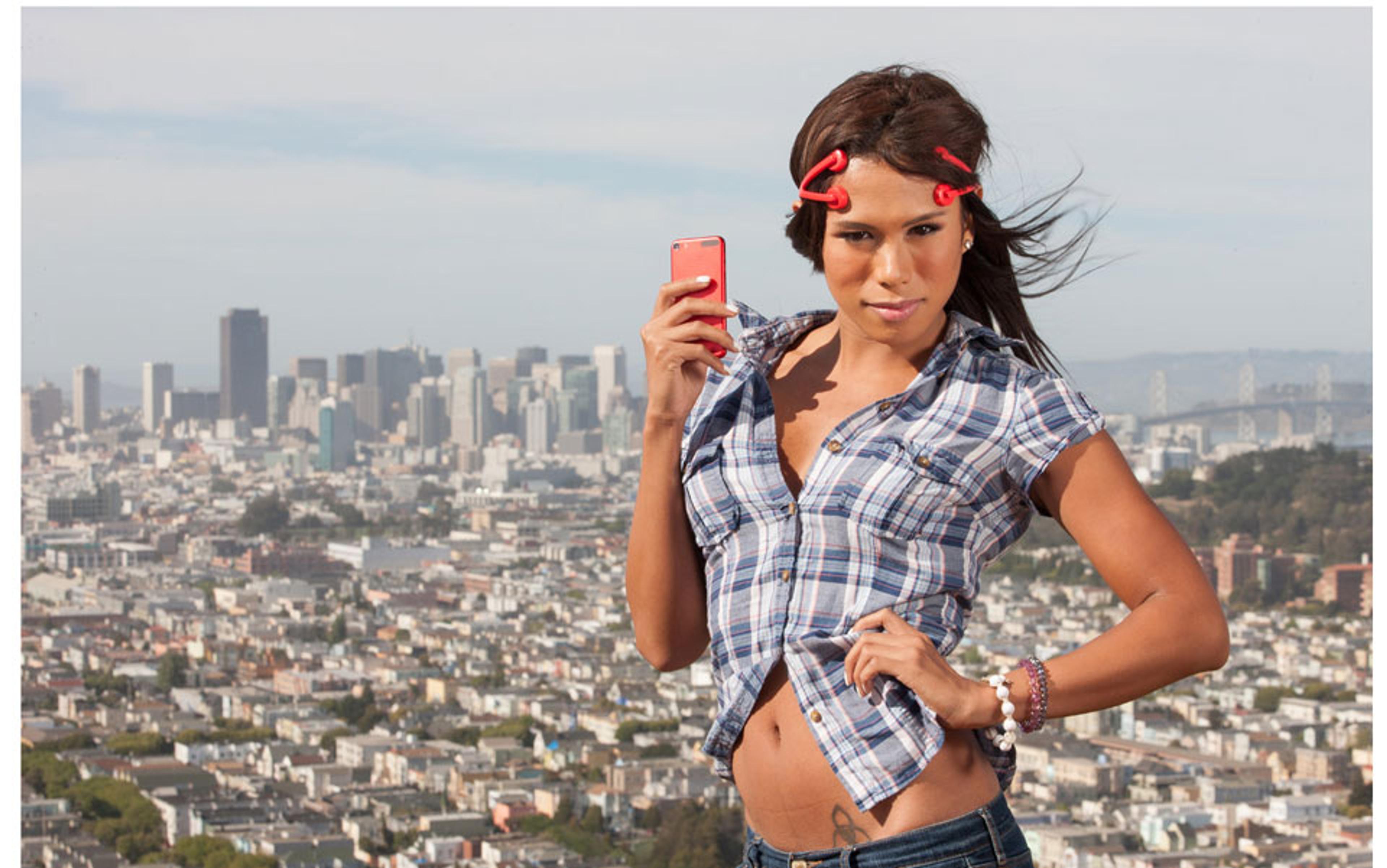 Photo of a woman with red curlers holding a red phone, wearing a plaid shirt, posing in front of a cityscape with buildings and a bridge.