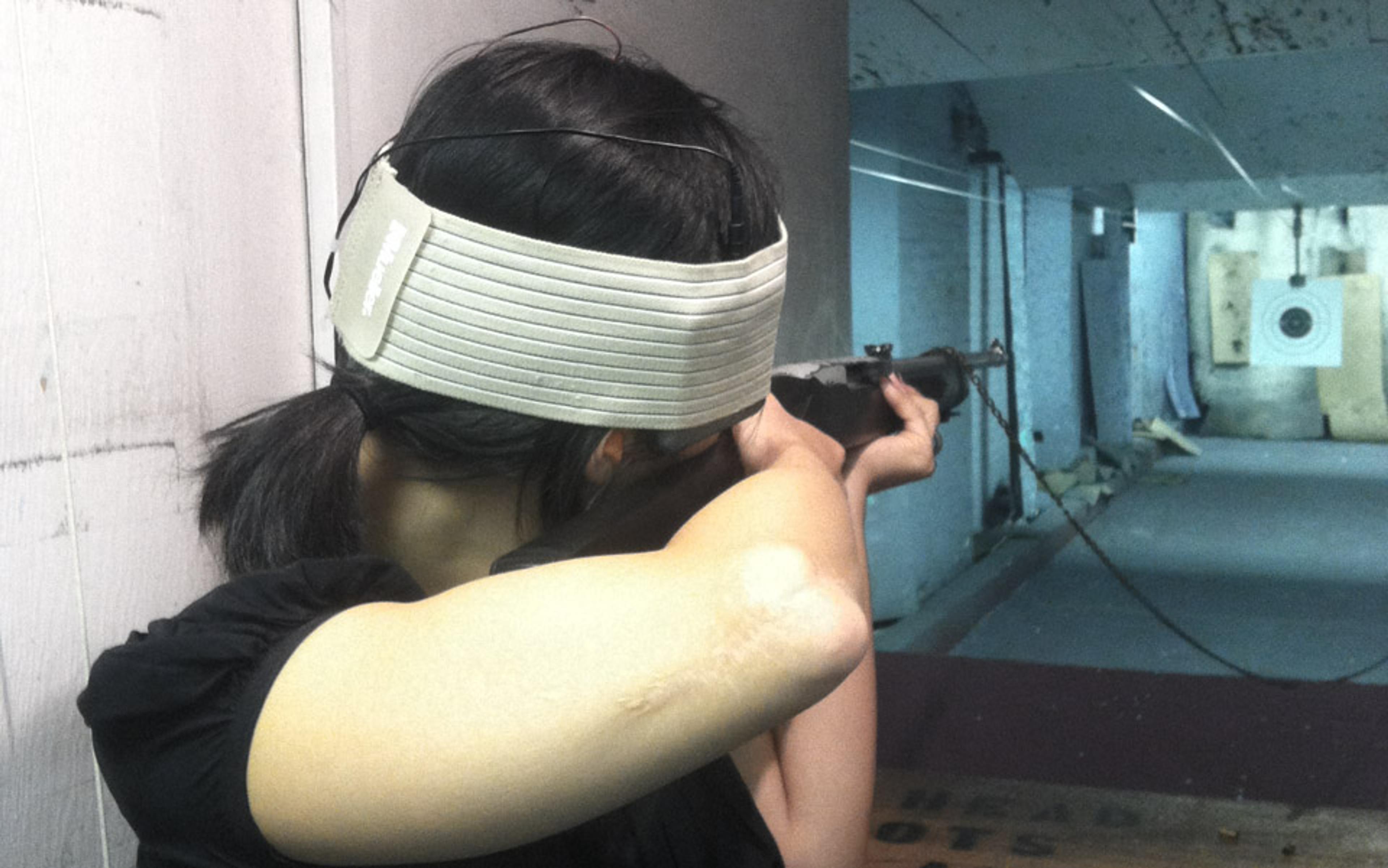 A woman with a pony tail aiming a rifle at a target inside an indoor shooting range wearing a head band.