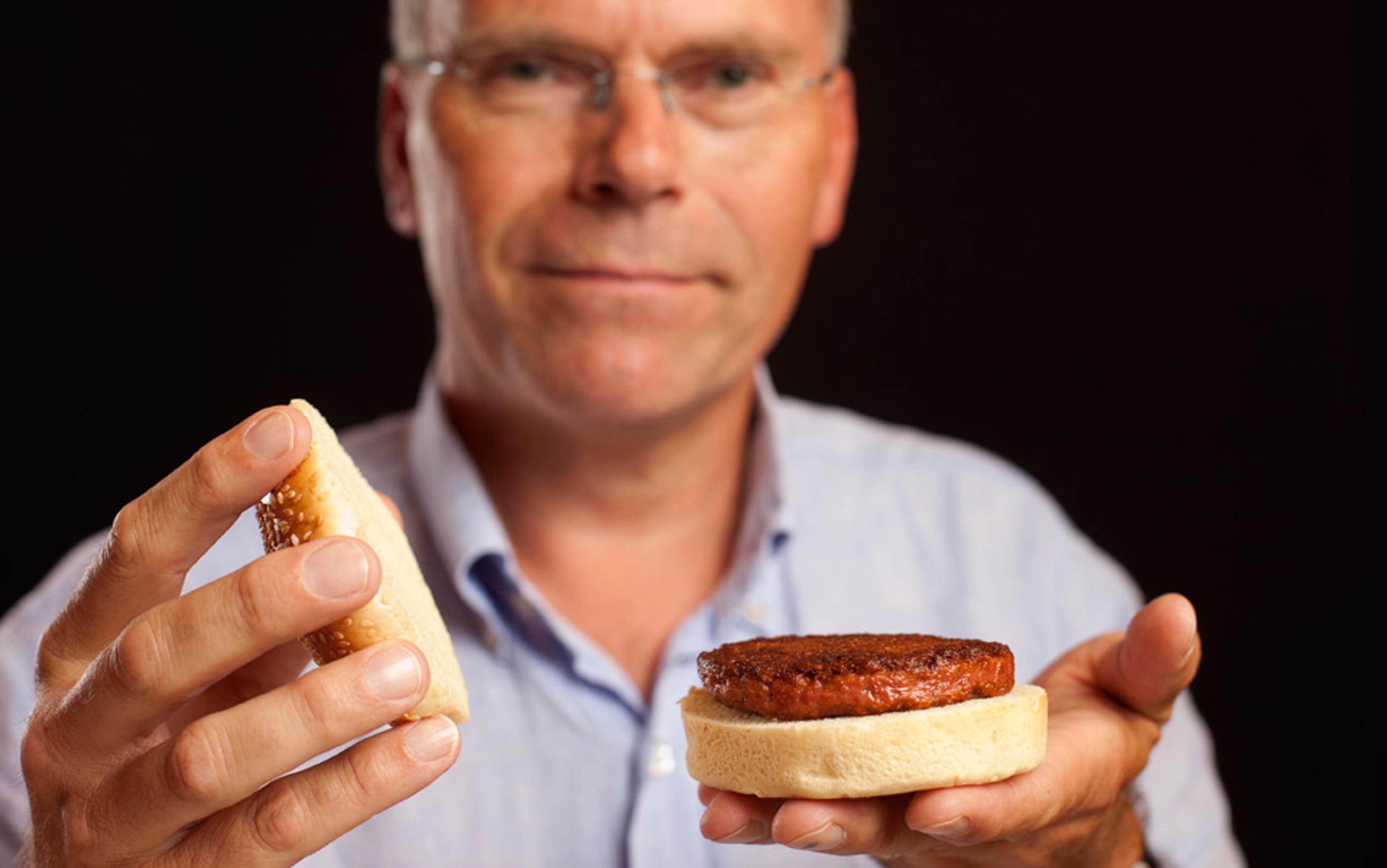 A man holding an open hamburger bun with a cooked burger patty on one half and the top bun in his other hand.